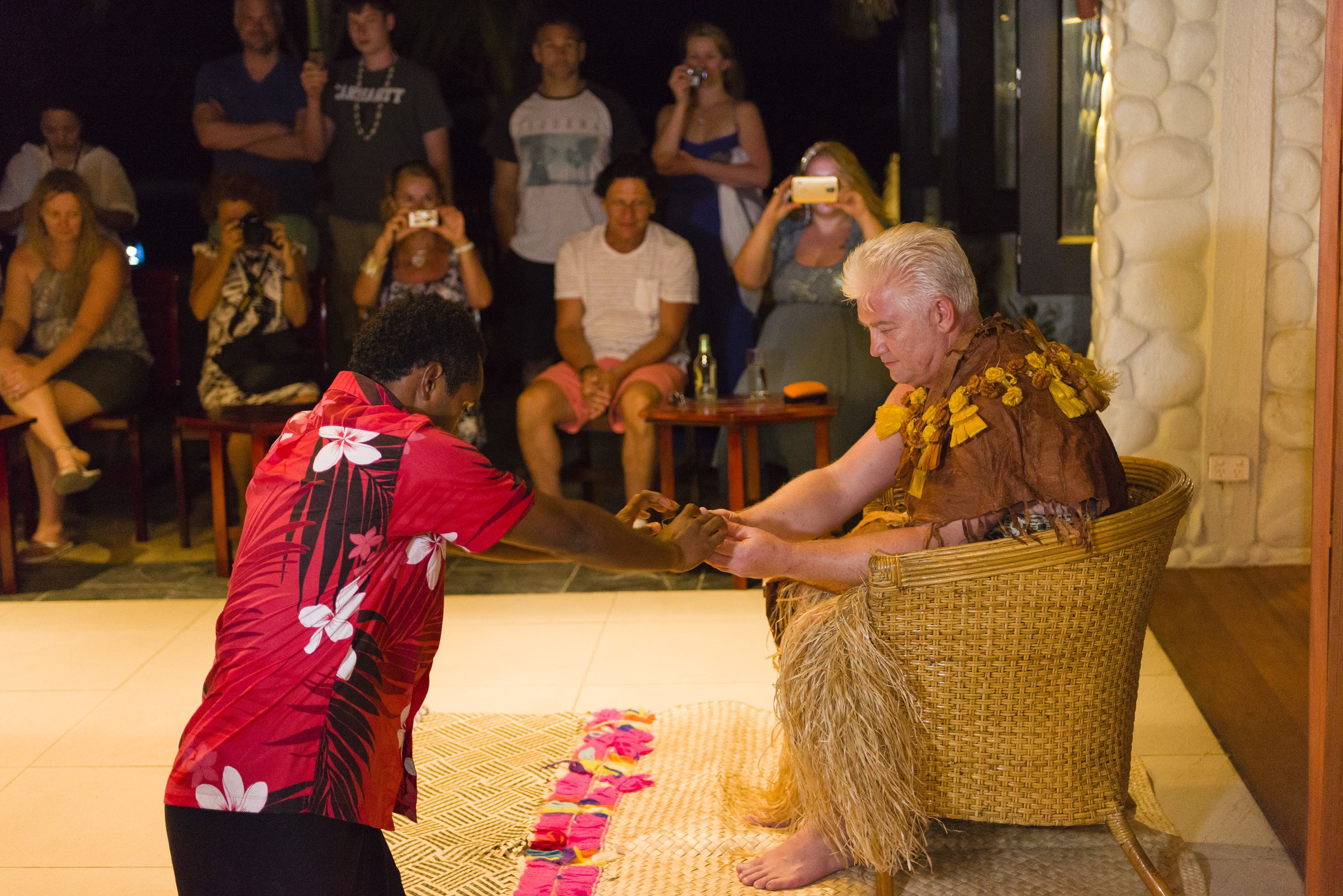 A person in a red floral shirt performing a traditional dance in front of an audience seated on chairs.