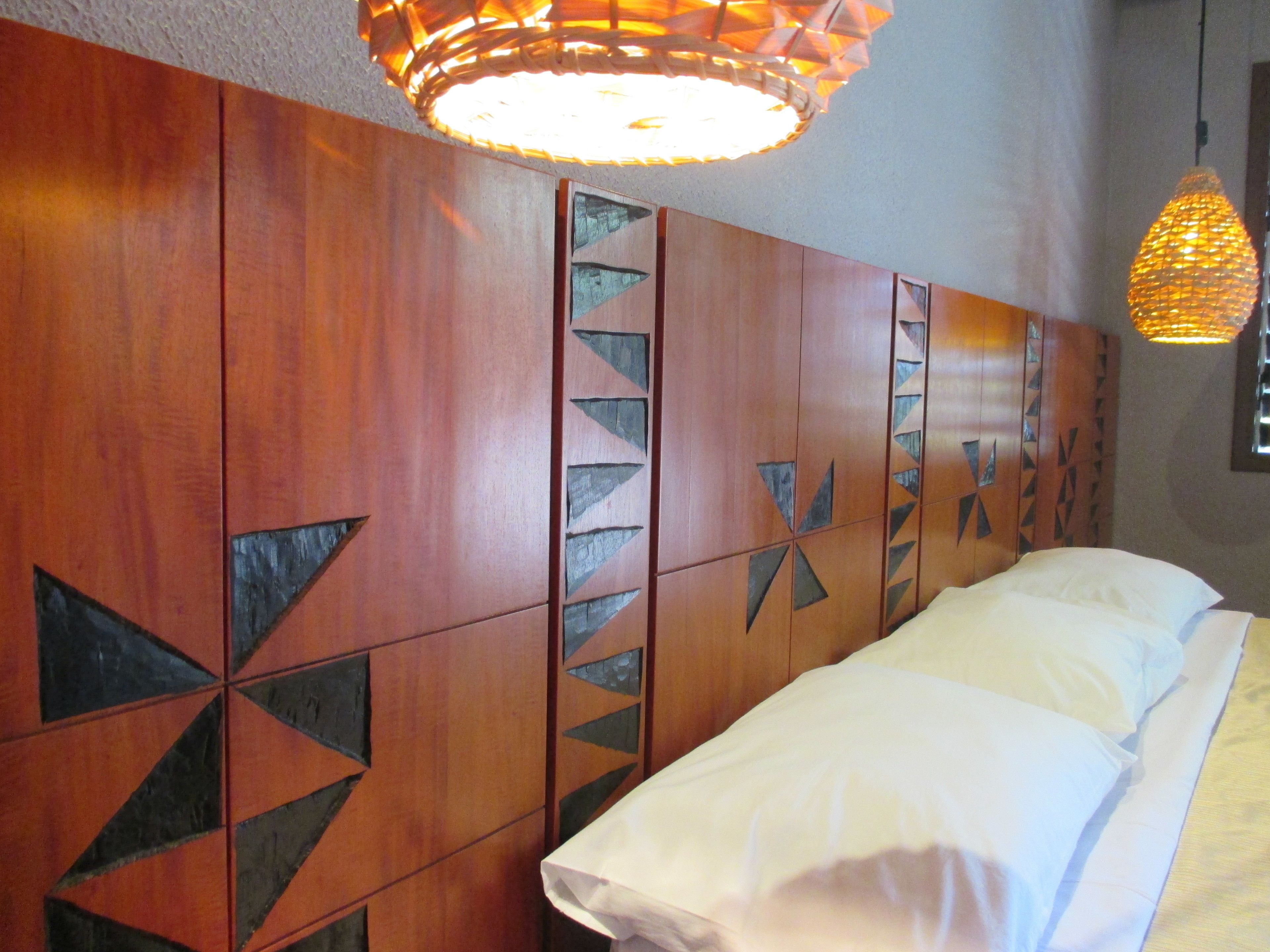 A room with a bed, wooden headboard with geometric patterns, and two hanging lamps.