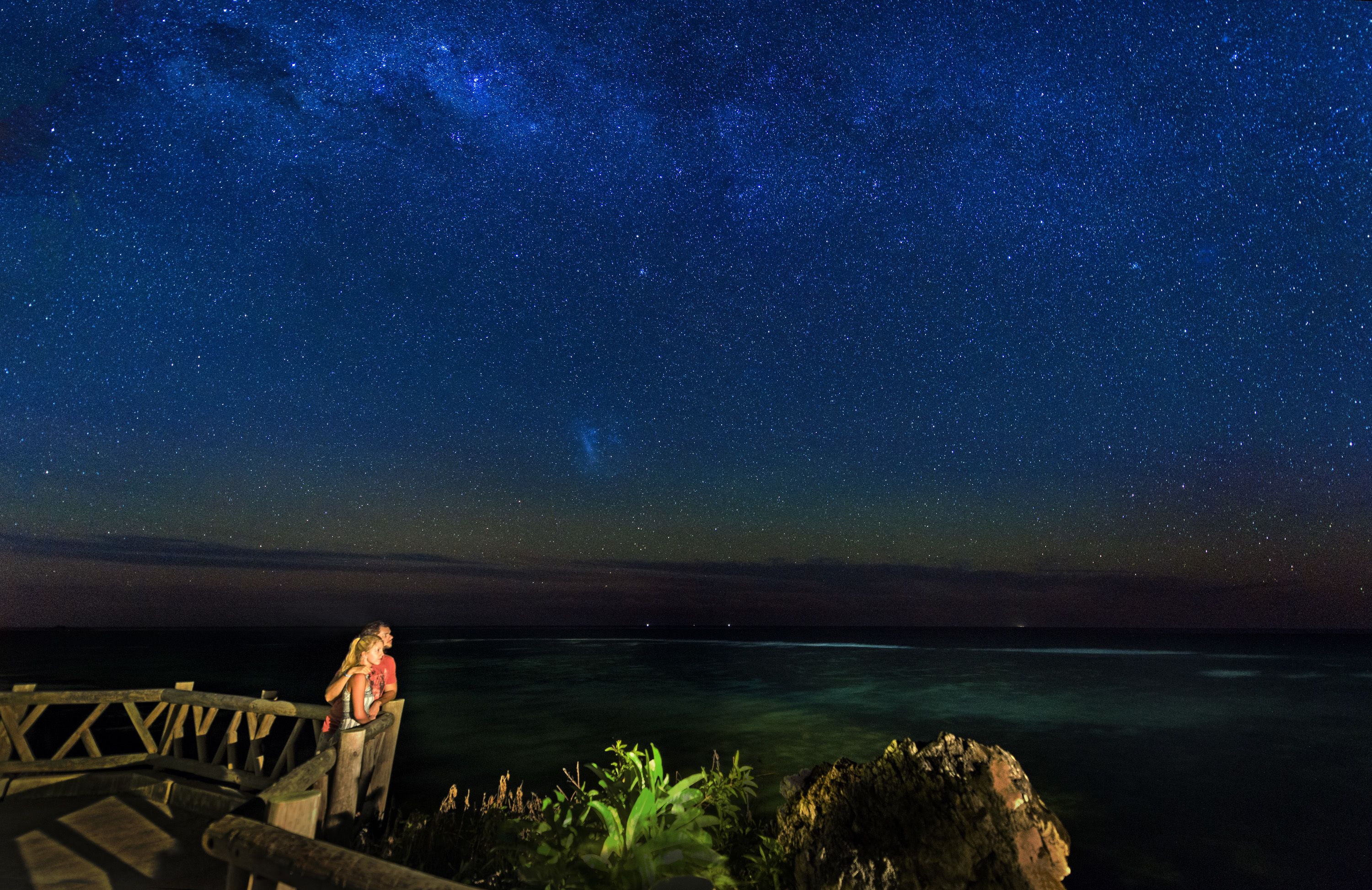 A night sky with stars and the Milky Way over a coastal area with a wooden viewing platform.