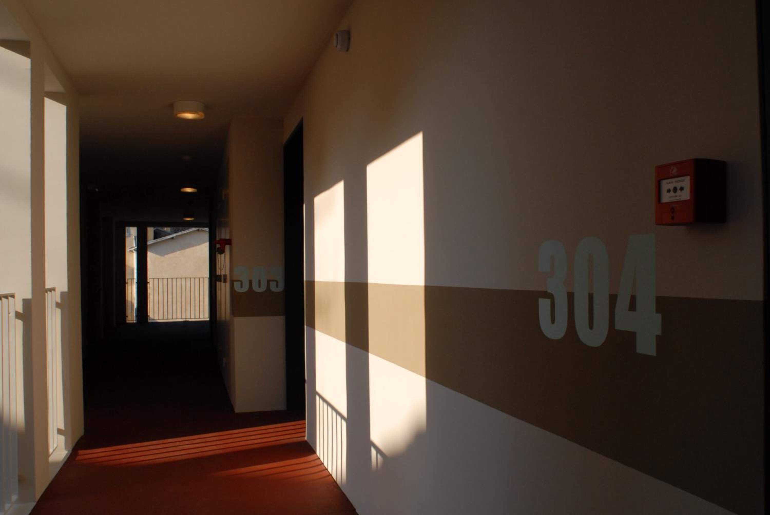 A hallway with a number 304 sign on the wall, a fire alarm, and a view of a building outside.