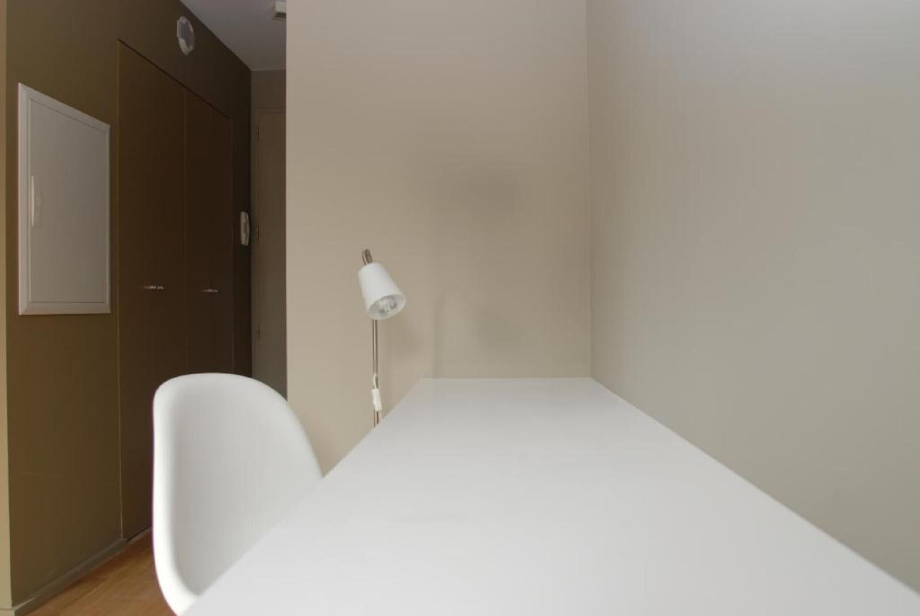 Een kleine, schone kamer met een wit bed, een witte stoel en een bedlampje.