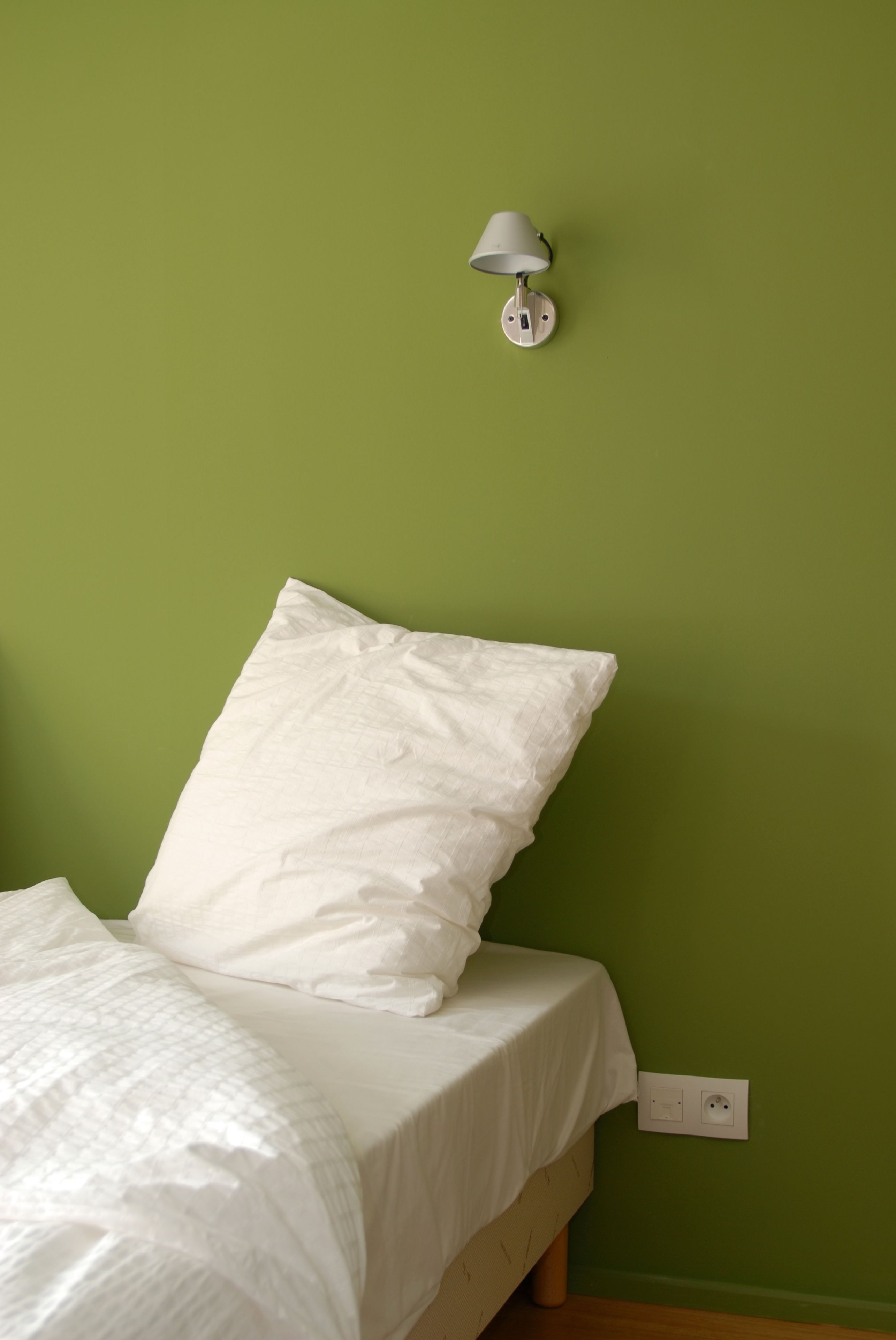 Un lit avec une literie blanche et un seul oreiller, adossé à un mur vert, une lampe fixée au mur et une prise de courant.