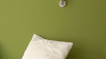 Un lit avec une literie blanche et un seul oreiller, adossé à un mur vert, une lampe fixée au mur et une prise de courant.