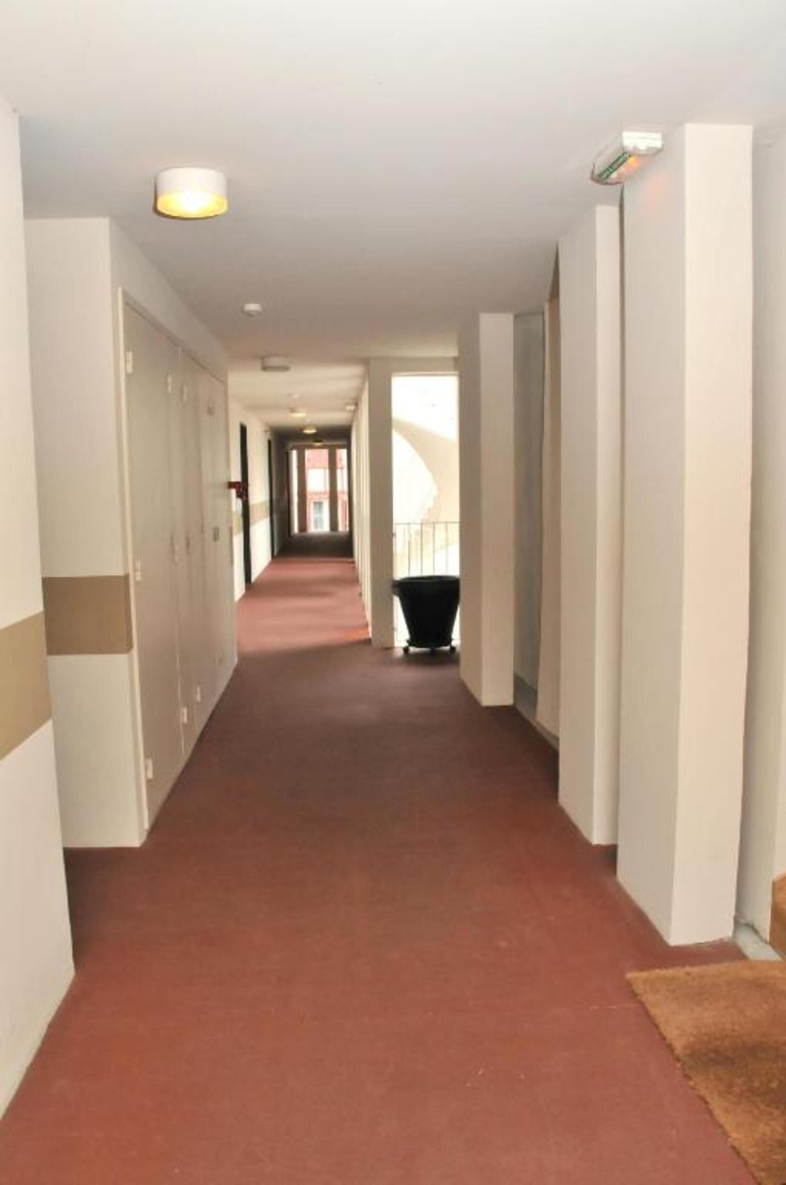 A long, narrow hallway with a red carpet, white walls, and a black trash bin at the end.