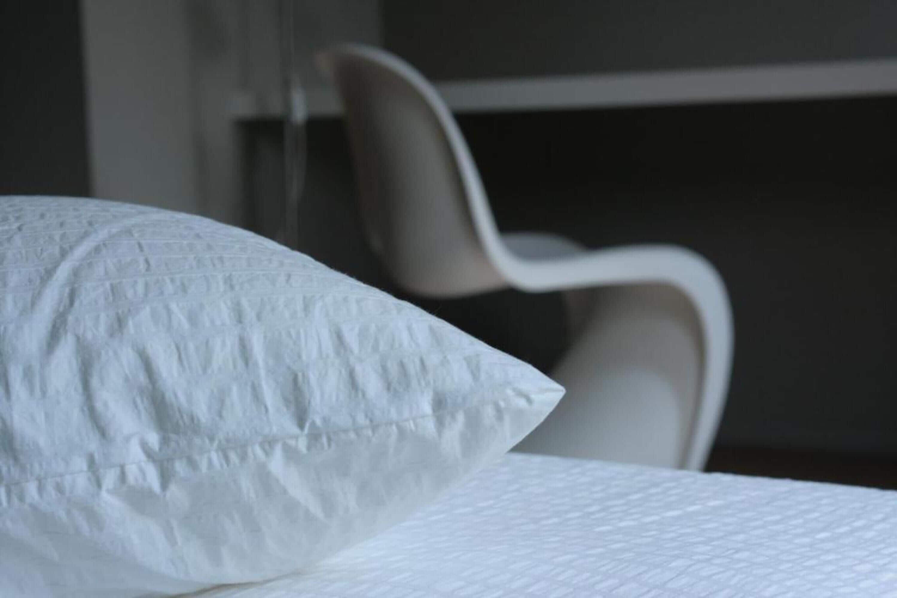 Una cama con un almohadón blanco y una silla al fondo.