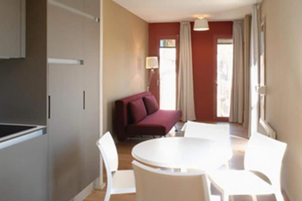 Un espace de vie compact comprenant un coin cuisine, un coin repas avec une table ronde et des chaises, un canapé rouge et une fenêtre avec des rideaux.