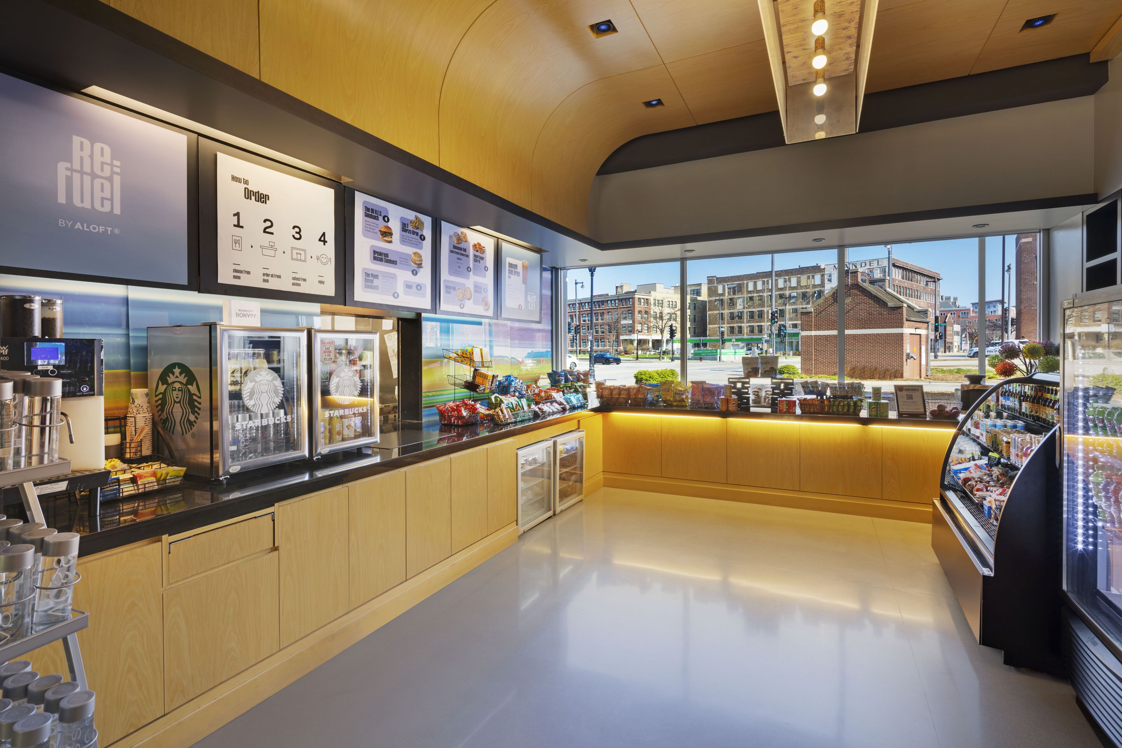 A modern coffee shop interior with a counter, Starbucks display, and a view of the city through large windows.