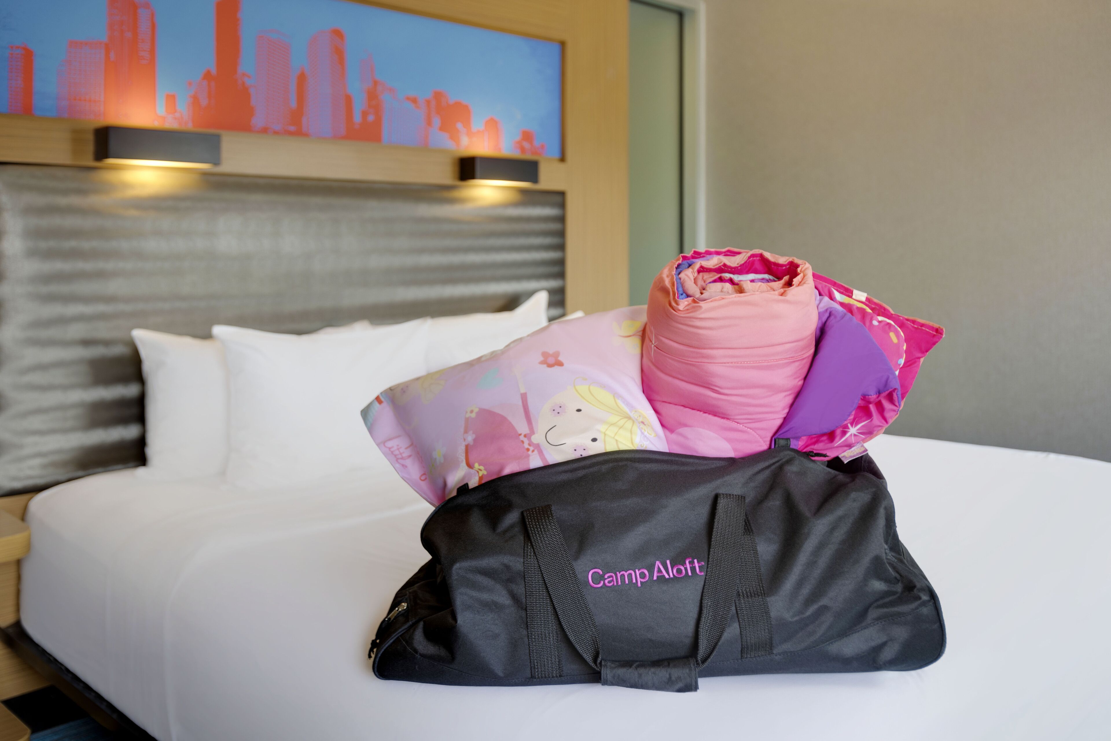 A bed with a black bag labeled "Camp ALoft" and a pink and purple bag with a floral pattern.