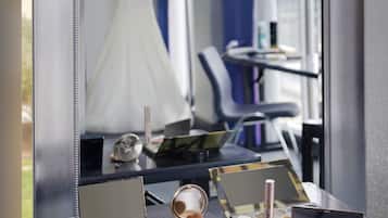 A vanity table with makeup and a mirror reflecting a room with a wedding dress and a chair.
