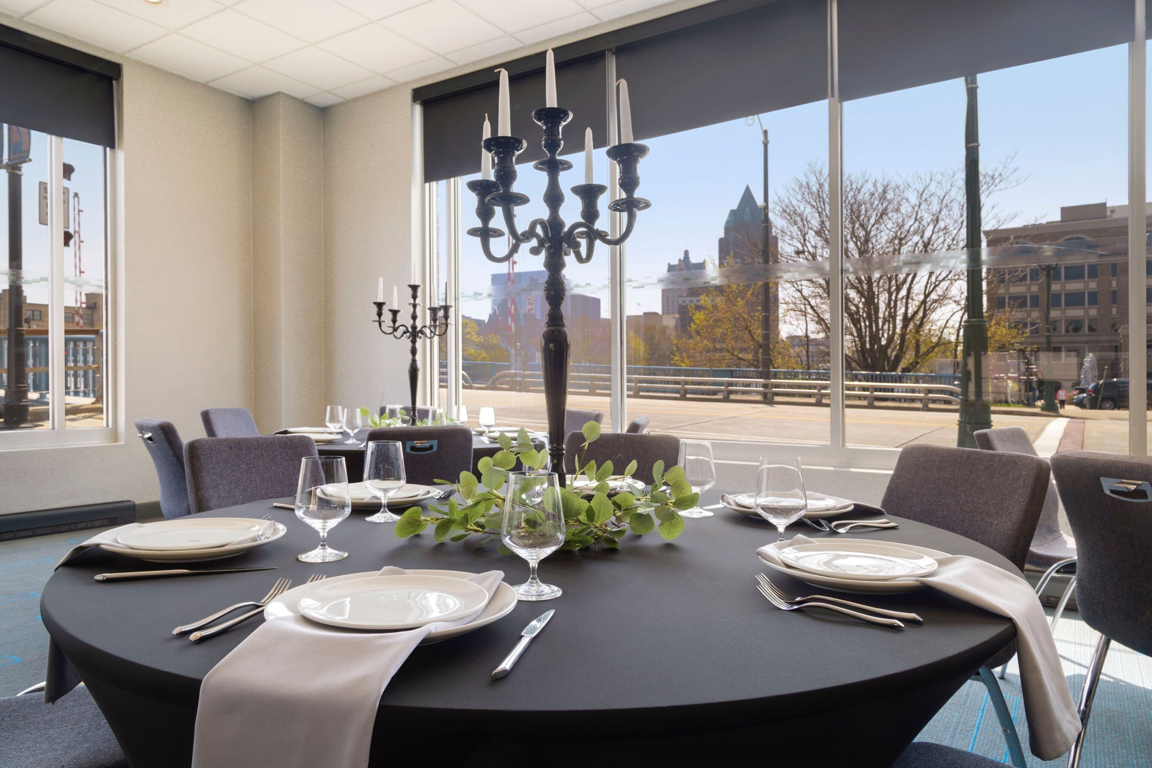 Une table à manger dressée pour six personnes, agrémentée d’un chandelier en guise de centre de table, de grandes fenêtres et d’une vue sur la ville.