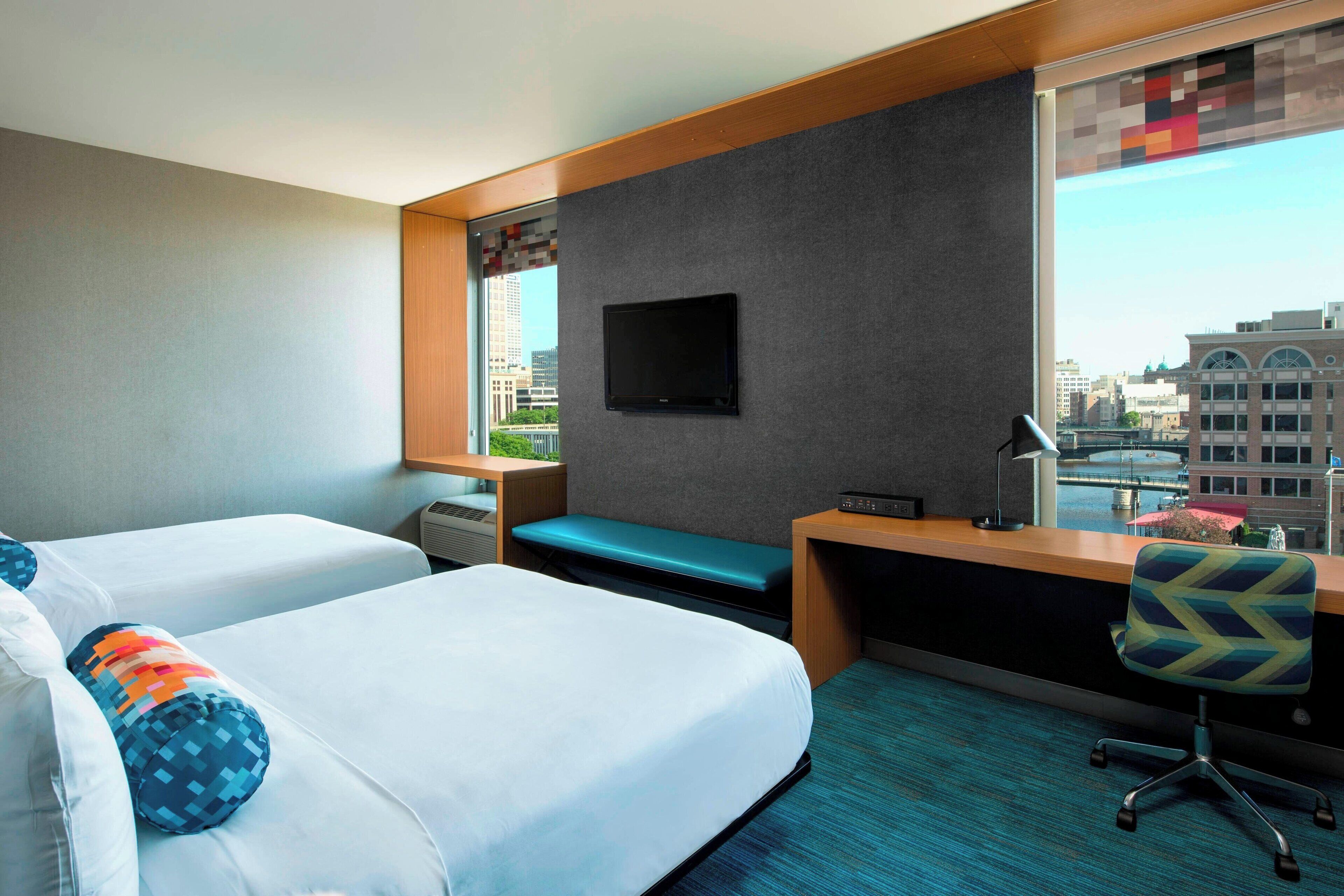 A hotel room with two beds, a desk, a chair, a TV, and a window with a city view.