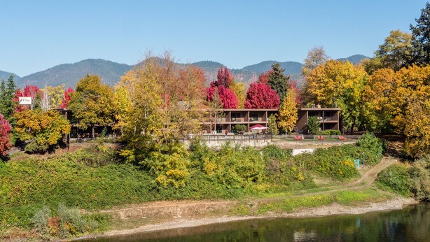 A hotel with a two-story structure, a covered outdoor seating area, and a view of a river and mountains in the background. - The Lodge at Riverside (Grants Pass)