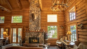 A log cabin interior with a stone fireplace, wooden walls, and large windows. - The Lodge at Riverside (Grants Pass)