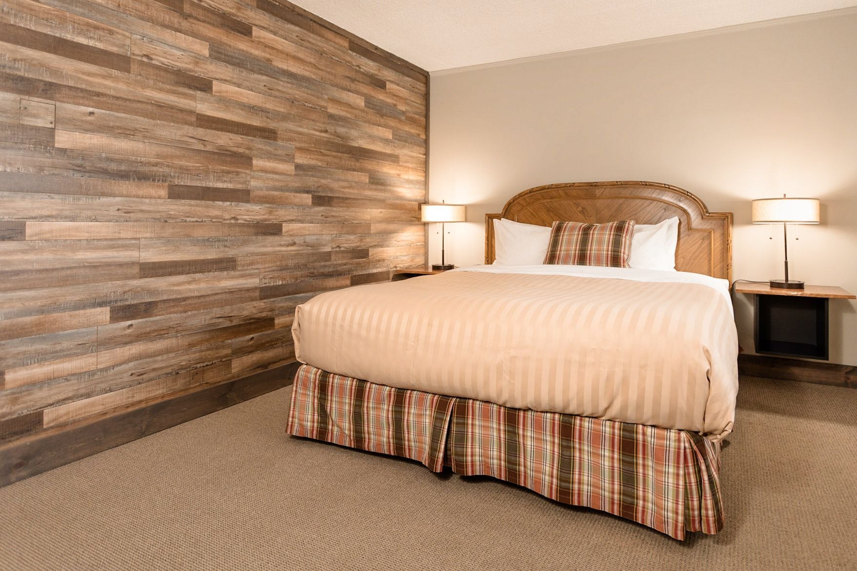 Een hotelkamer met een wand van houten panelen, een groot bed met een geruite sprei, twee nachtkastjes met lampen, en een hoofdeinde.