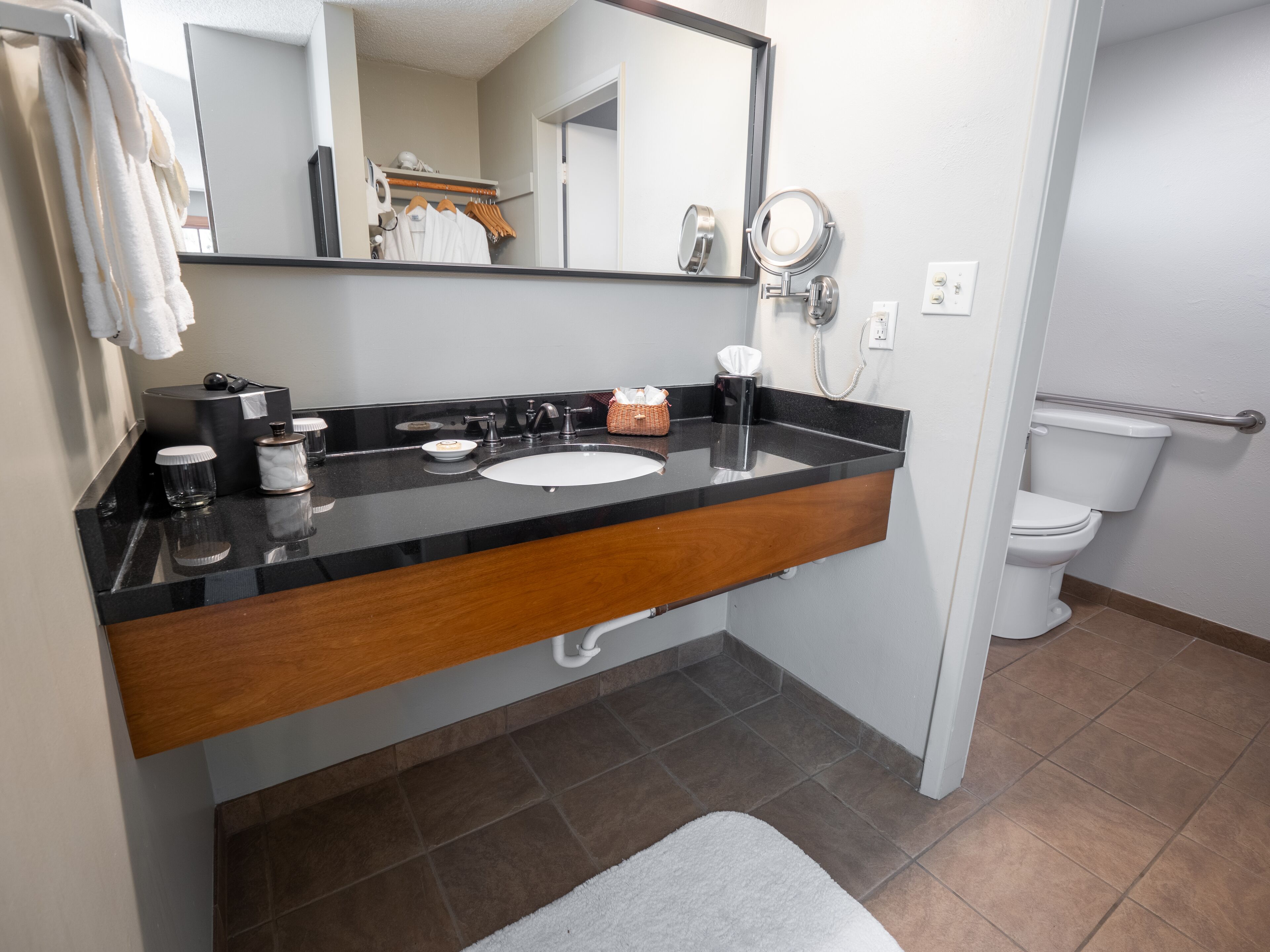 A bathroom with a black countertop, white sink, and a mirror. There is a towel rack, a basket, and a toilet visible.
