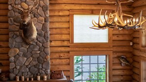 A log cabin interior with a stone wall, a mounted elk head, a chandelier, and a window with a view. - The Lodge at Riverside (Grants Pass)