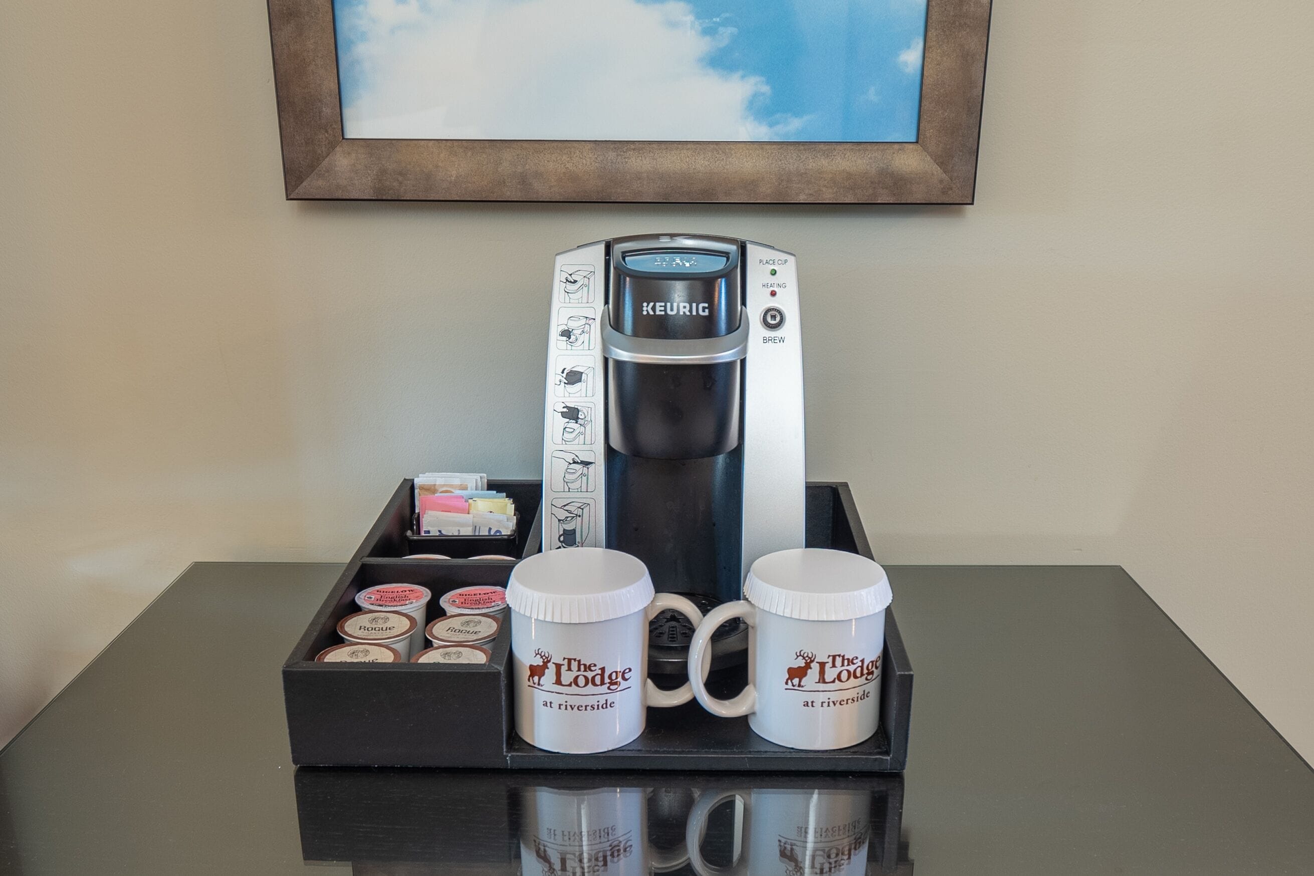Une machine à café dotée d’un panneau de commande, deux tasses portant le logo « The Lodge at Riverside », et un plateau contenant des dosettes de café et une petite boîte.