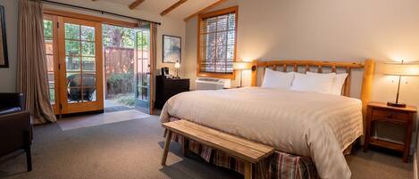 A bedroom with a large bed, wooden headboard, bedside lamps, a bench, a desk, and a view through a glass door to an outdoor area.