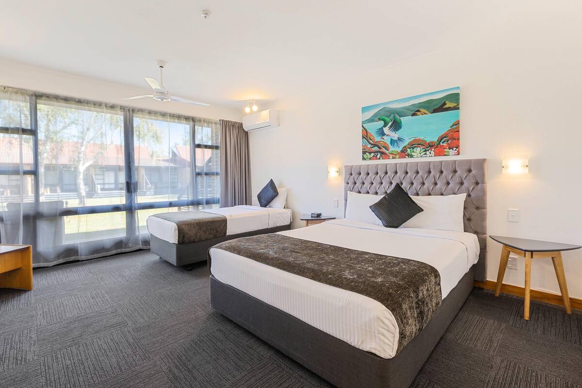 A hotel room with two beds, a large window, a painting, and a small table.