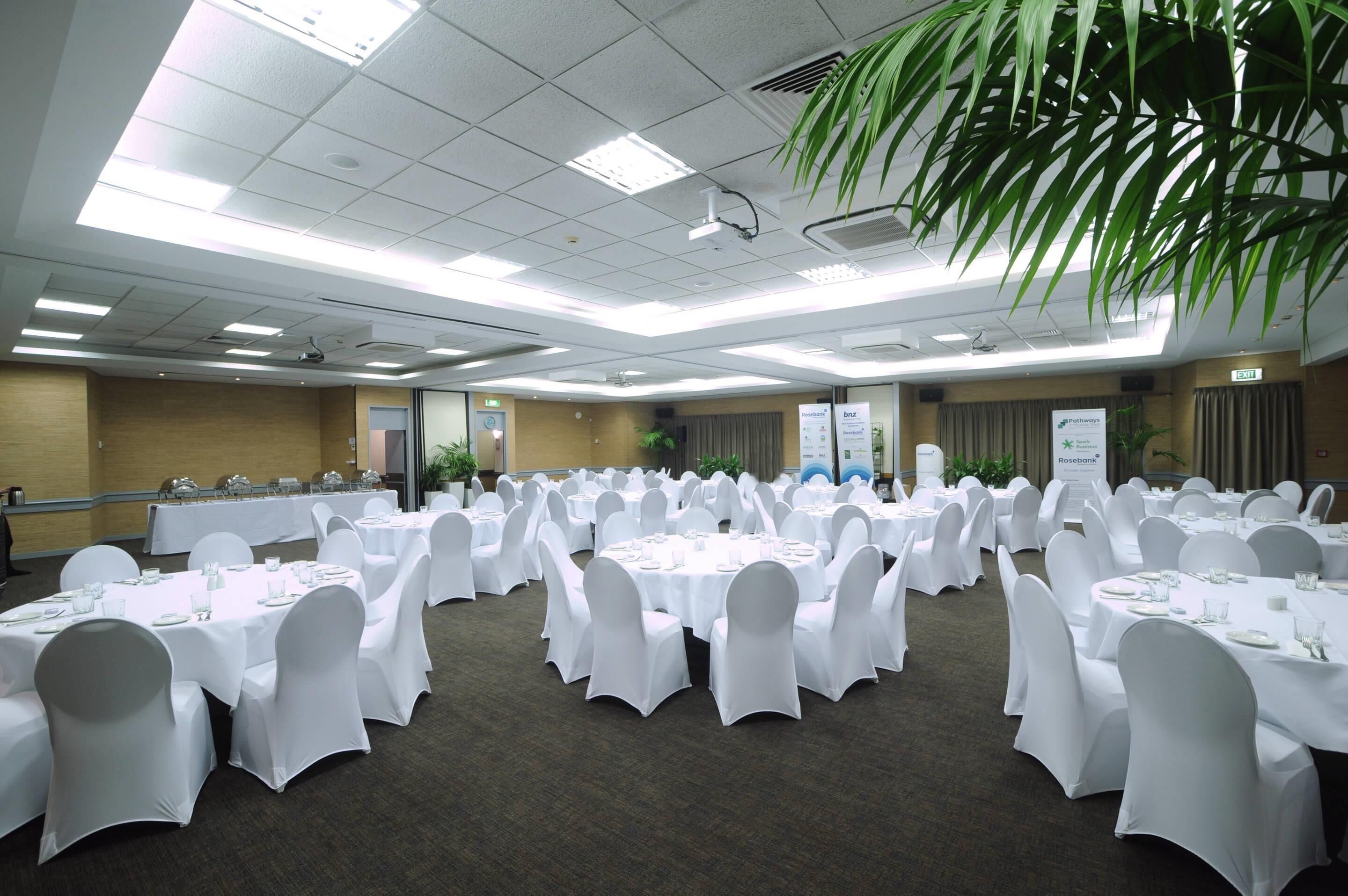 A conference room with multiple round tables, white tablecloths, and chairs, equipped with ceiling lights and a projector.