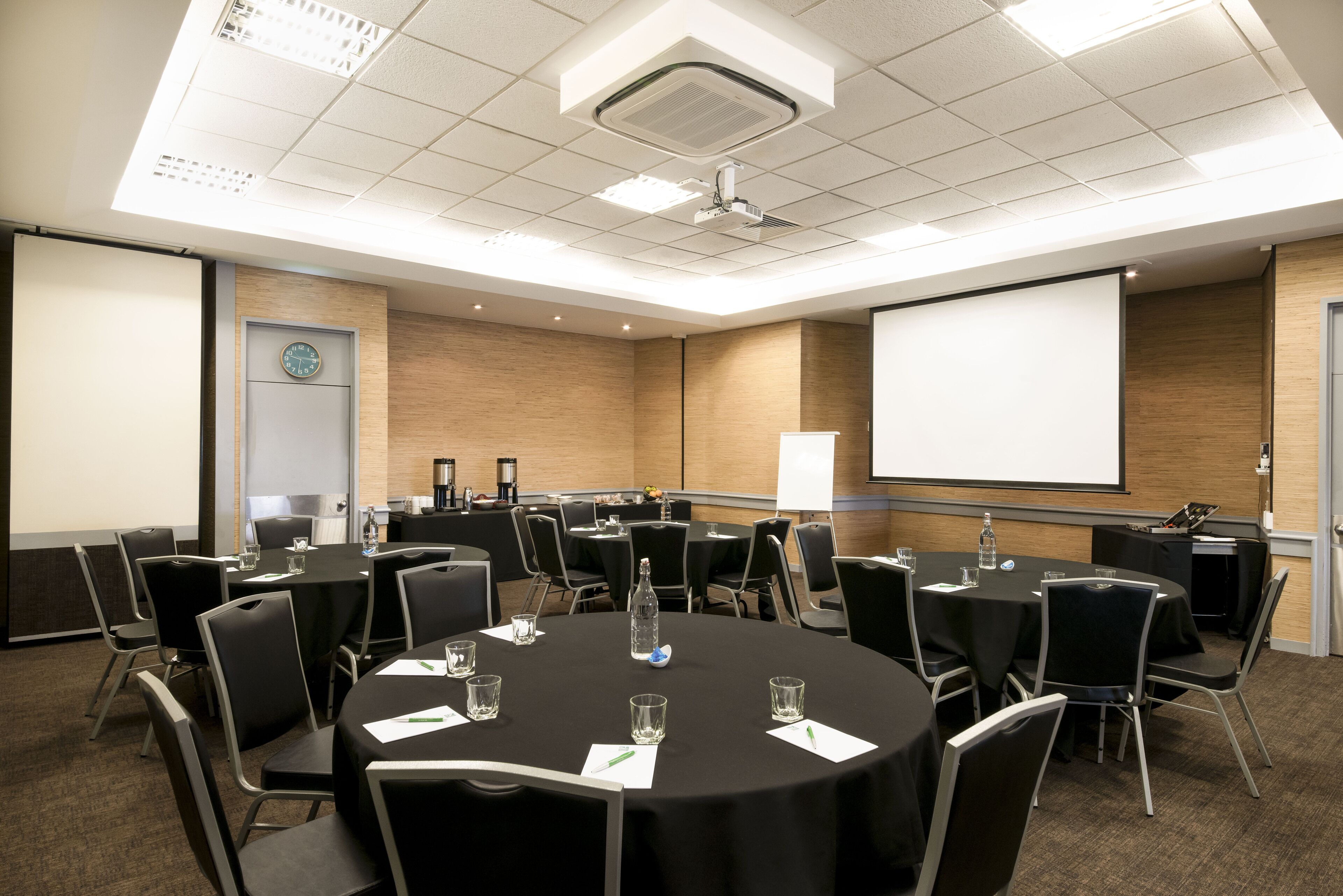 A conference room with multiple tables, chairs, a projector screen, and a clock on the wall.