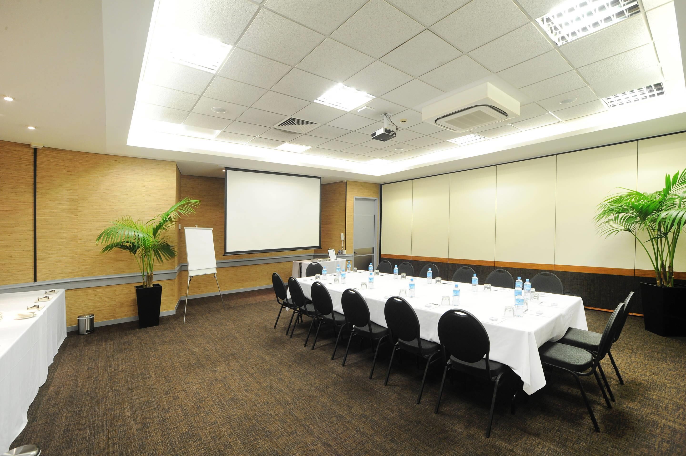 A conference room with a long table, chairs, a projector screen, and two potted plants.