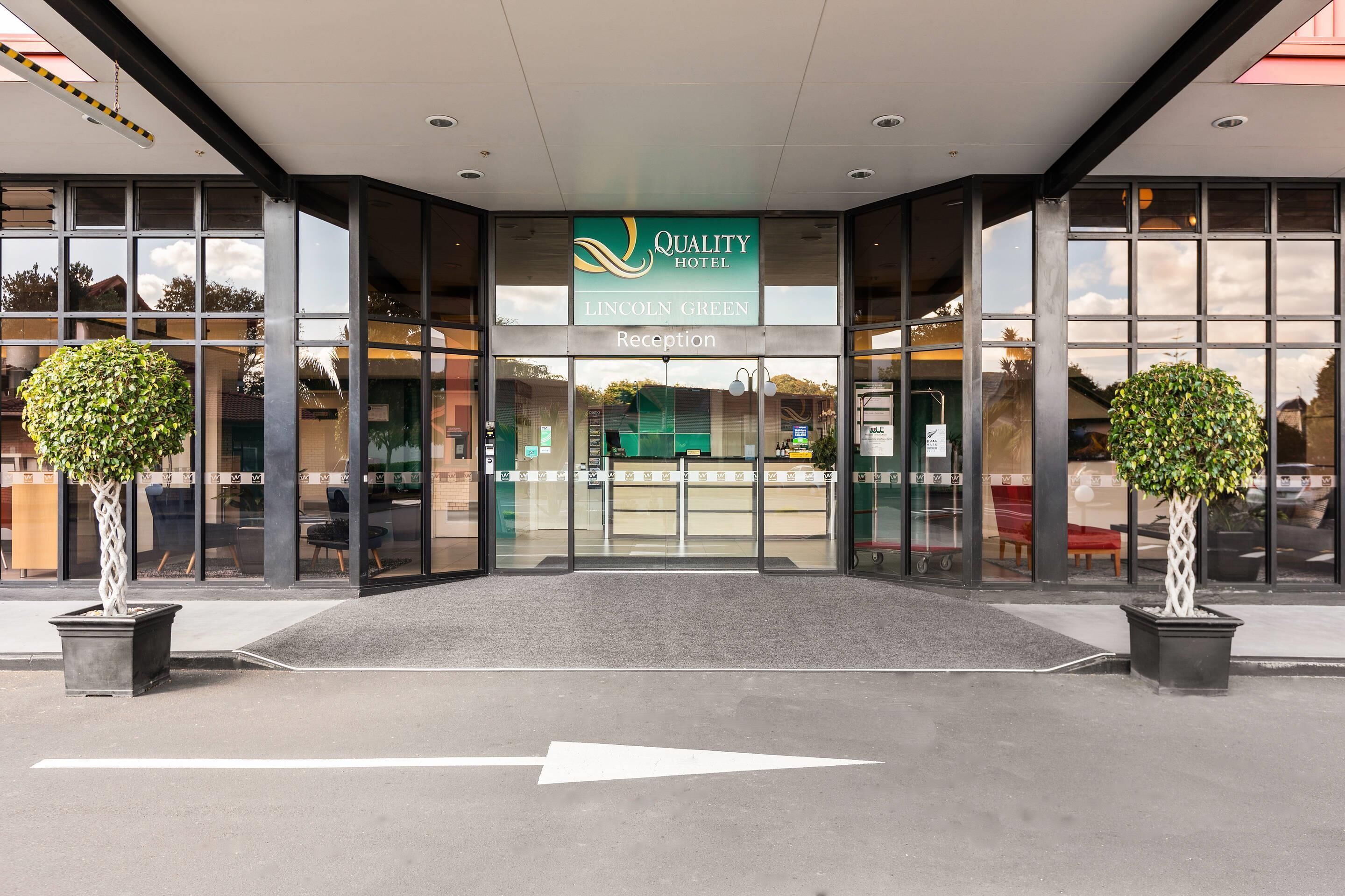 A hotel entrance with a reception desk, glass doors, and two potted trees.