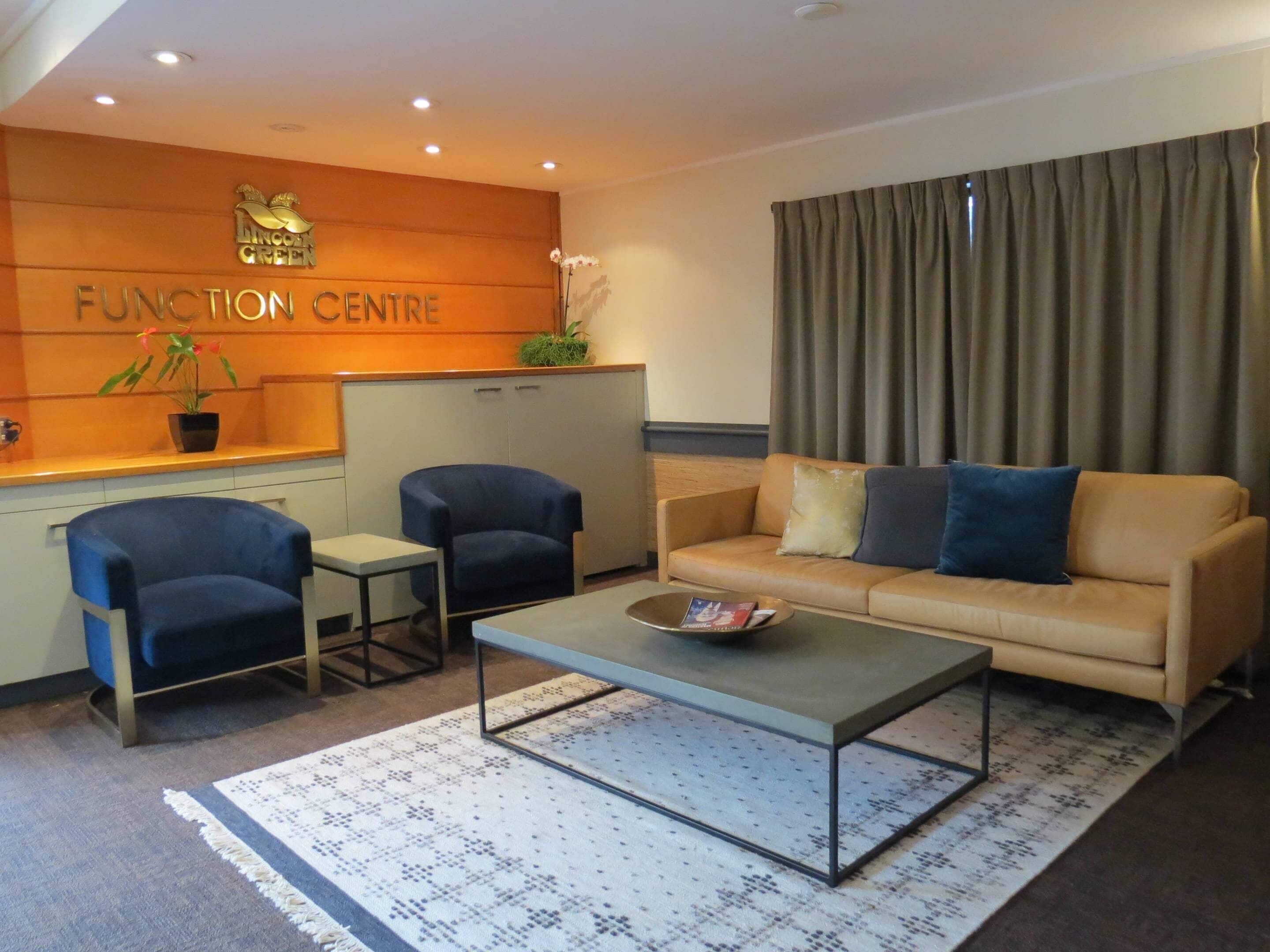 Una sala de estar moderna con un sofá, dos sillones, una mesa de centro y un mostrador de recepción con un cartel que dice 'FUNCTION CENTRE'.