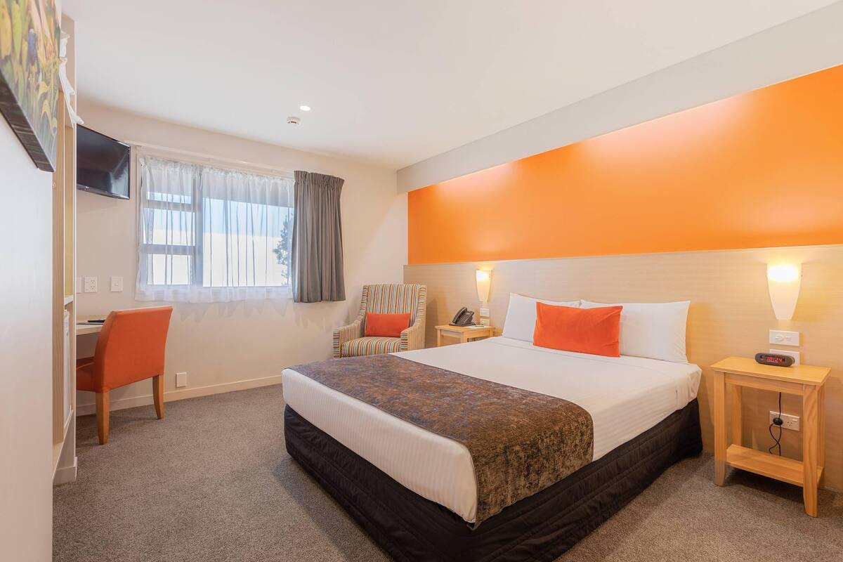 A hotel room with a large bed, a desk, a chair, a TV, and a window with curtains.