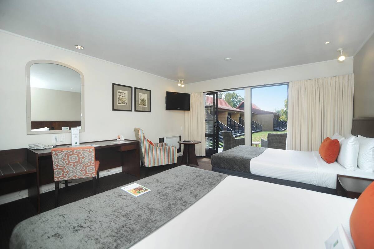 A hotel room with a desk, two beds, a chair, a TV, and a view of the outdoors.