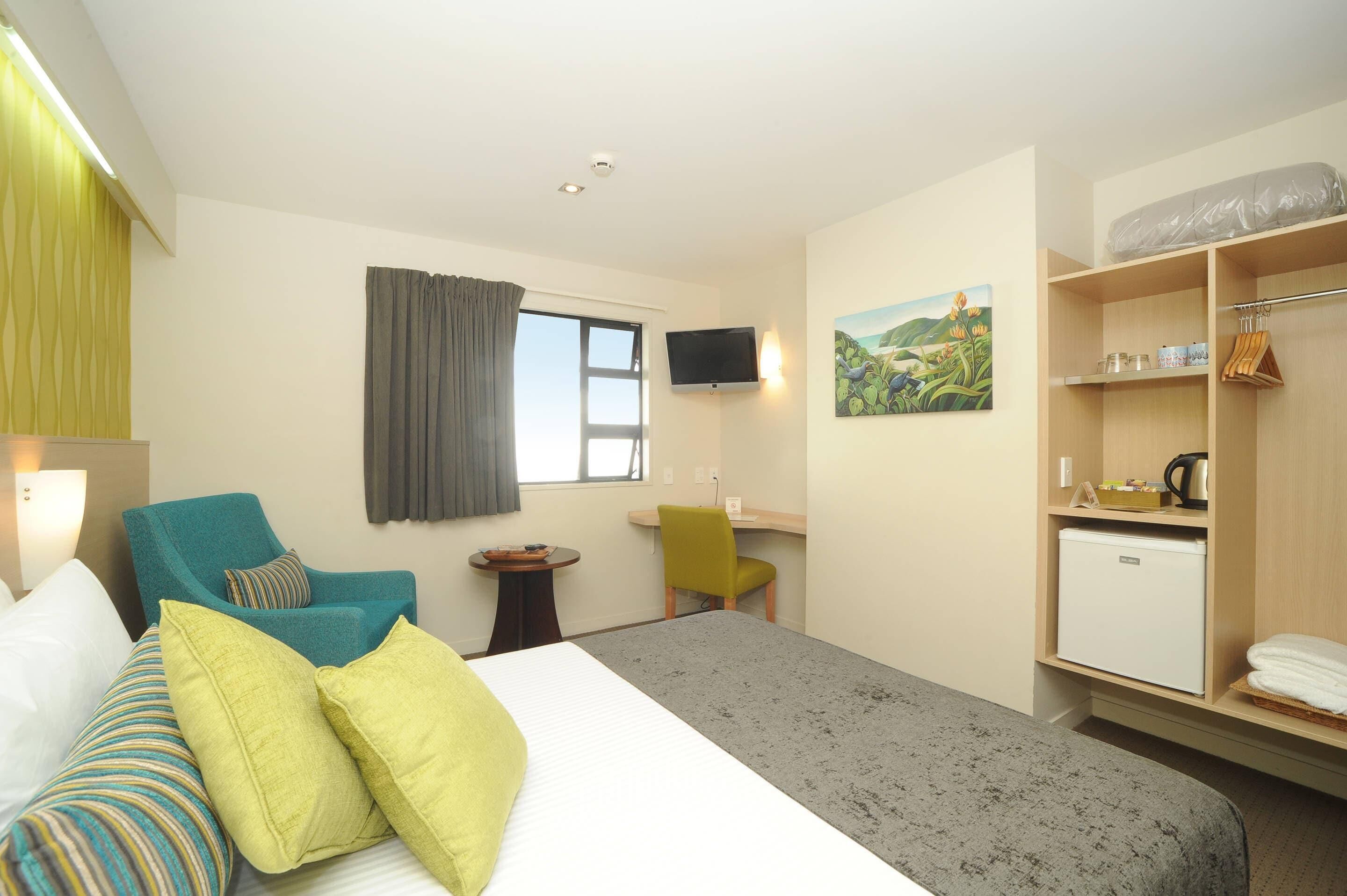 Quarto de hotel com cama, duas cadeiras, escrivaninha, televisão, pintura e mini-geladeira.