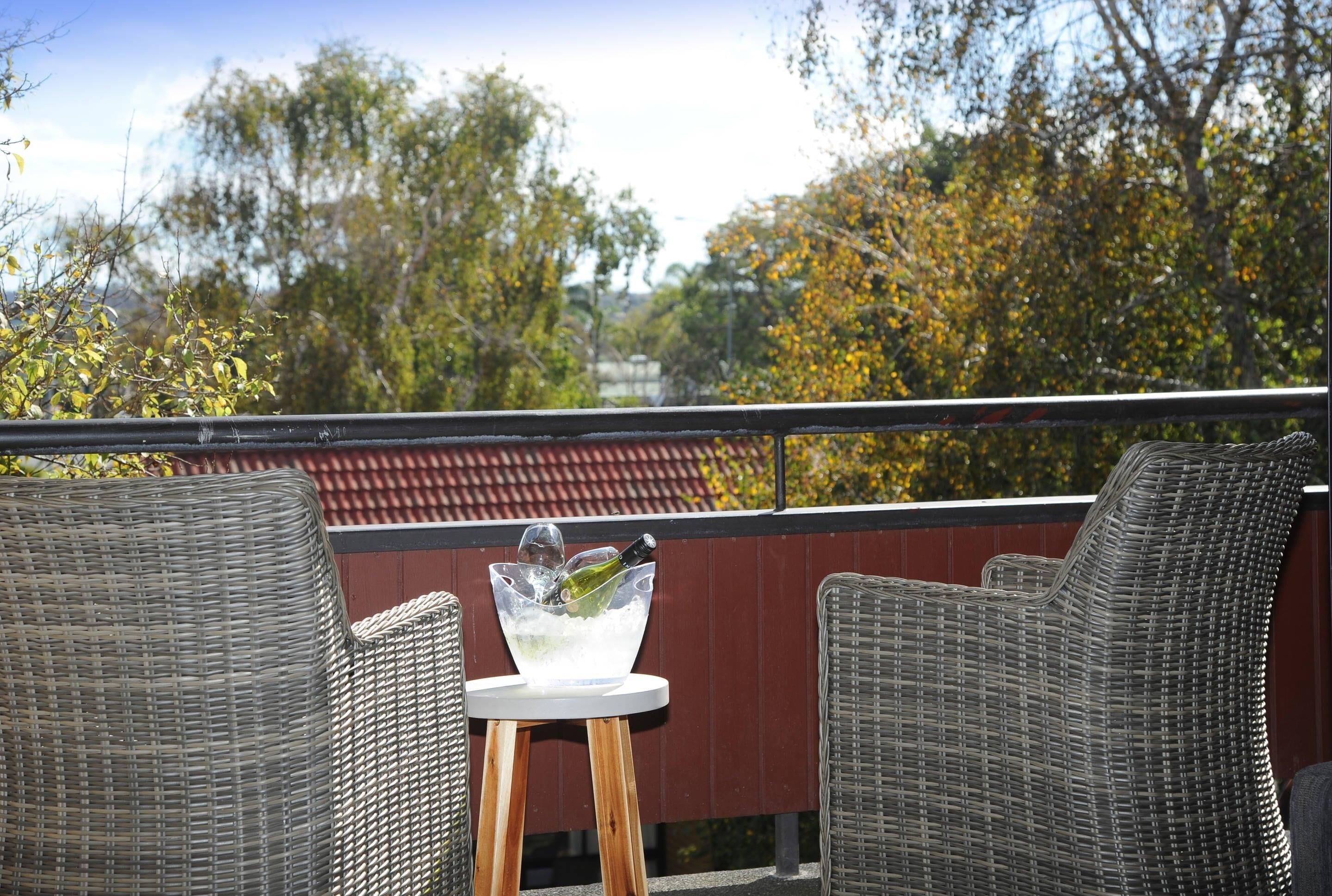 Une chaise et une table en osier, avec un saladier en verre contenant une bouteille et des glaçons, sur un balcon.