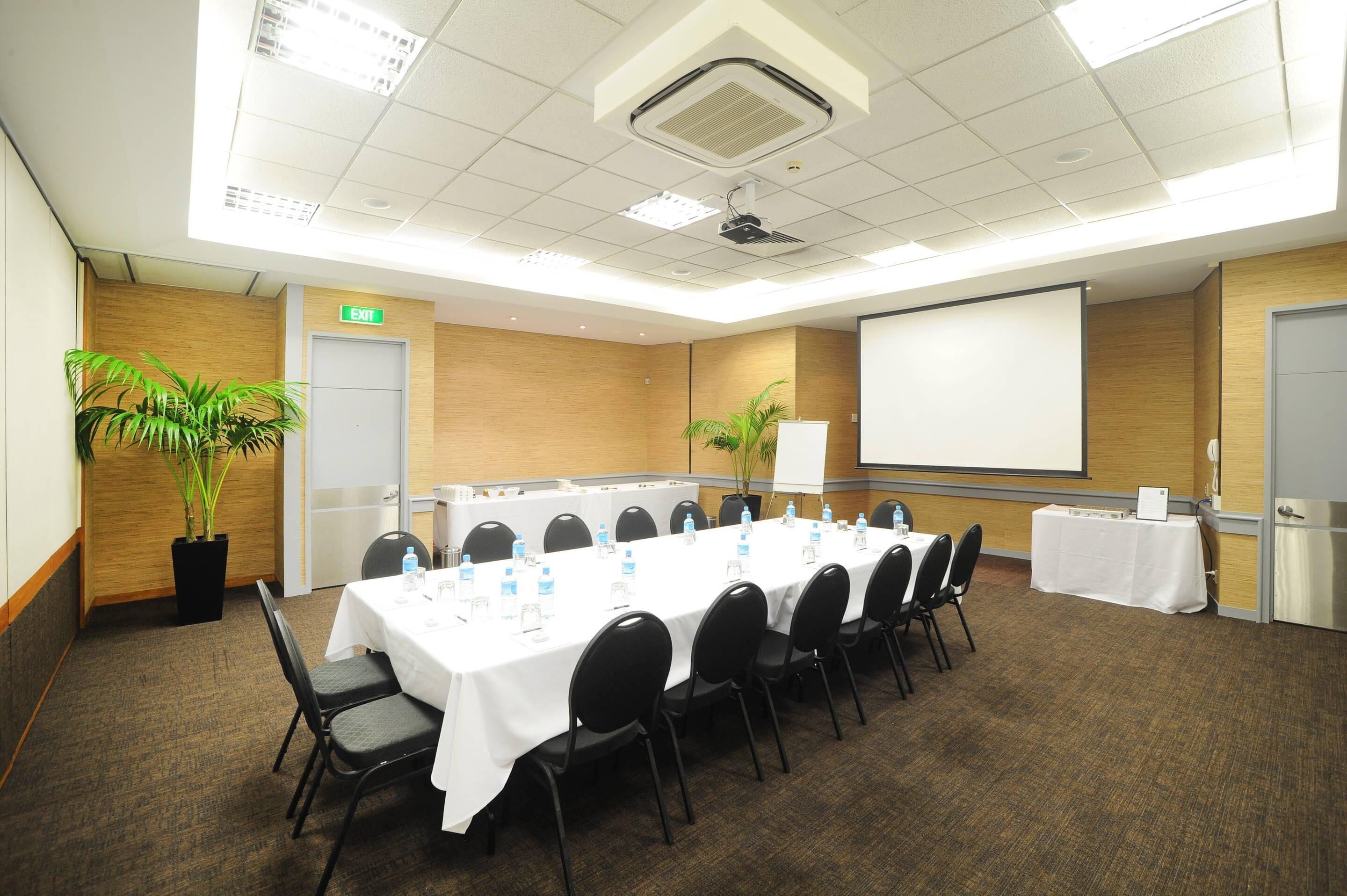 Ein Konferenzraum mit einem langen Tisch, Stühlen, einer Projektionsleinwand und zwei Zimmerpflanzen.