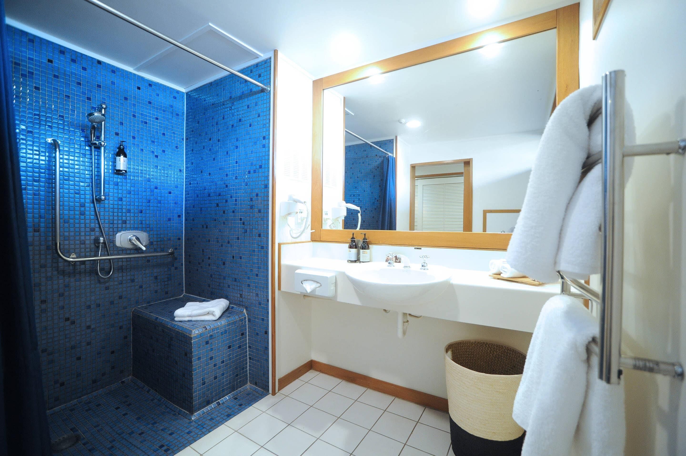 A bathroom with a blue-tiled shower, a white sink, a mirror, and a towel rack.