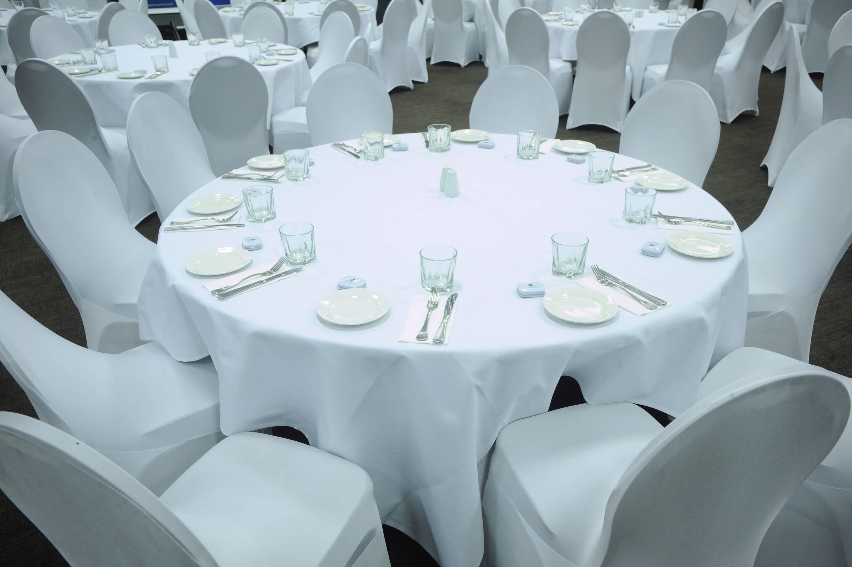 A round table with white tablecloths and chairs, surrounded by other similarly arranged tables and chairs.