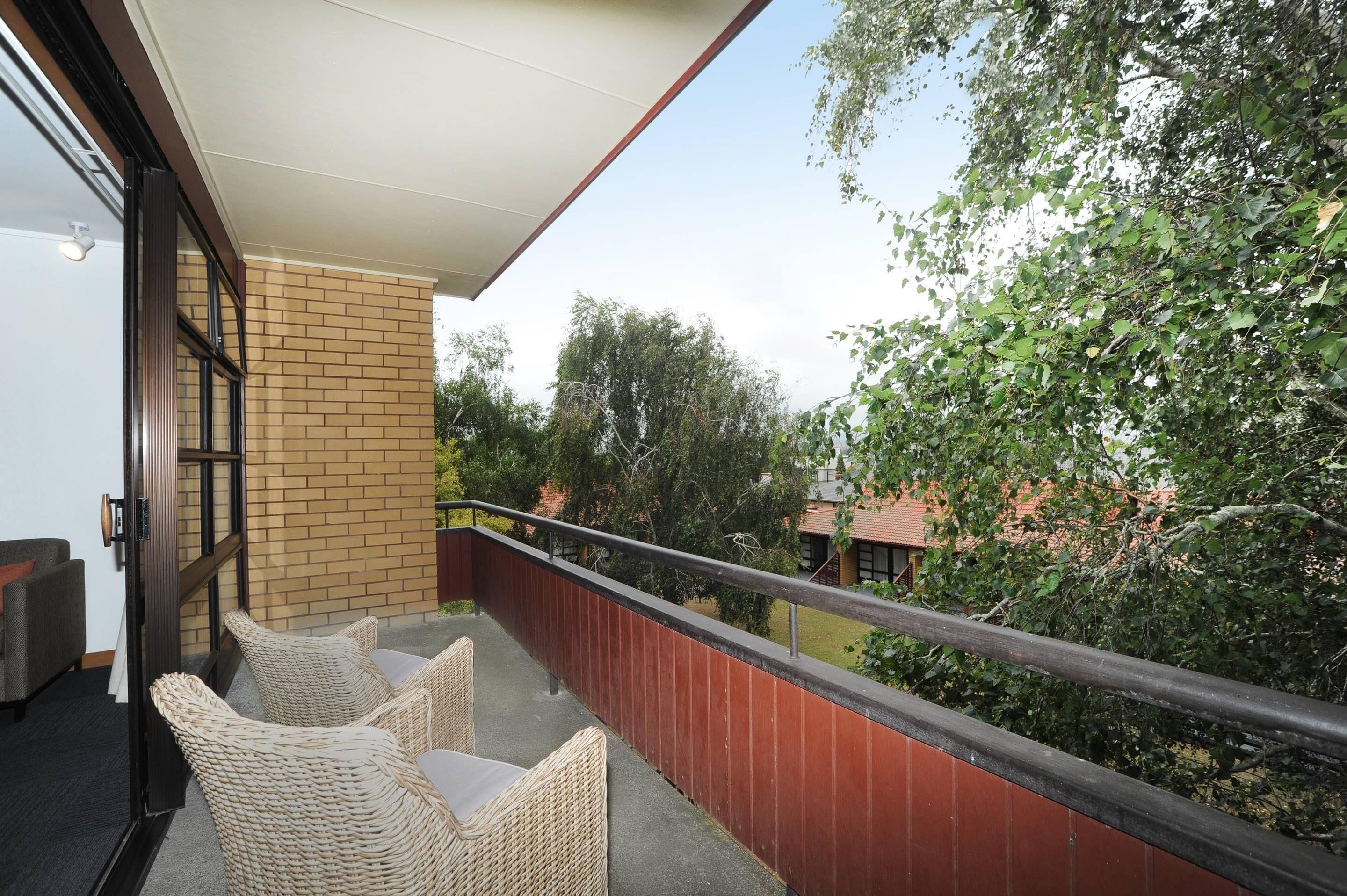 A balcony with wicker furniture, a brick wall, and a view of trees and a building in the background.