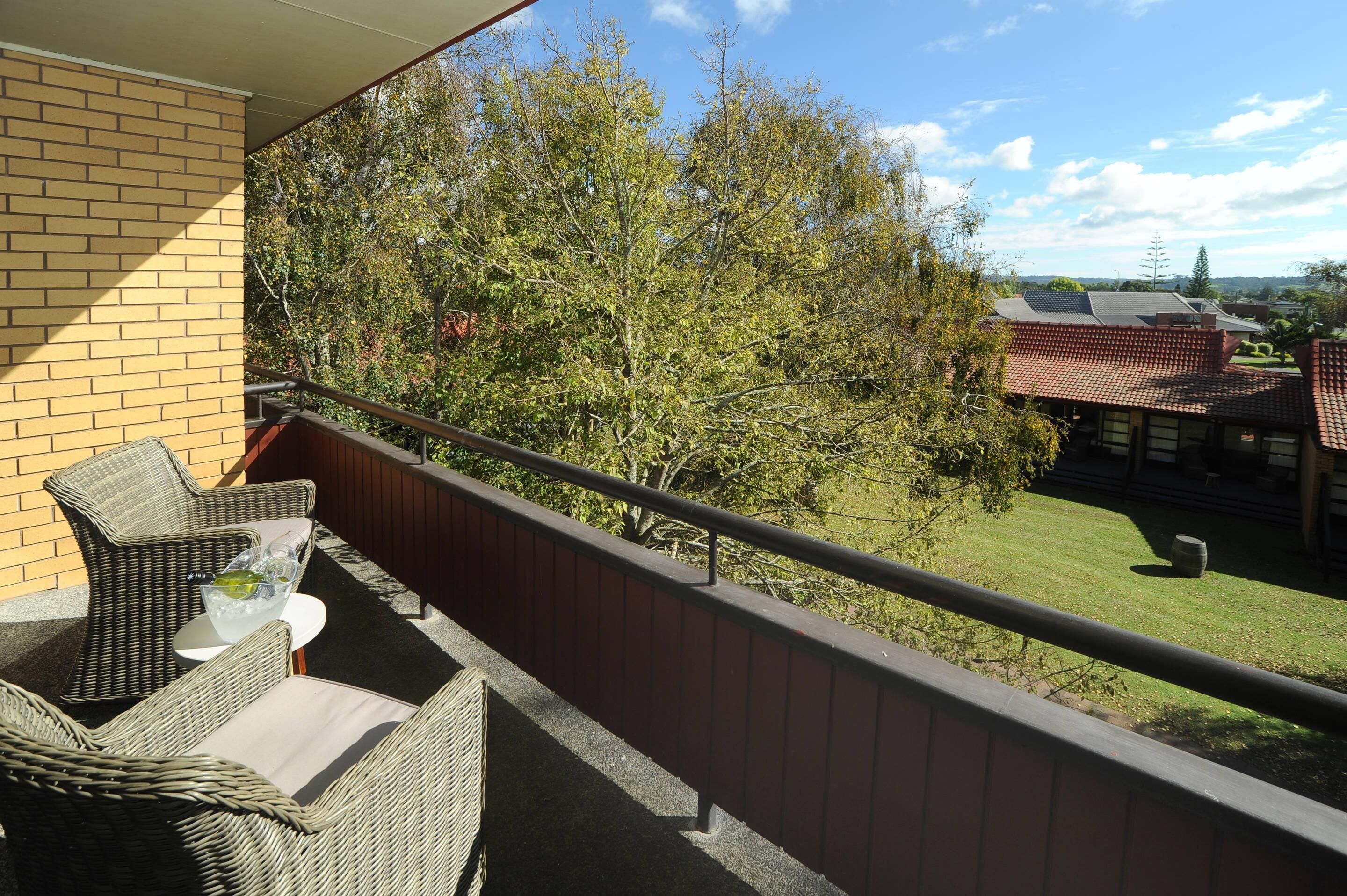Un balcón con mobiliario de mimbre, una mesa de vidrio y vistas a árboles y una casa.