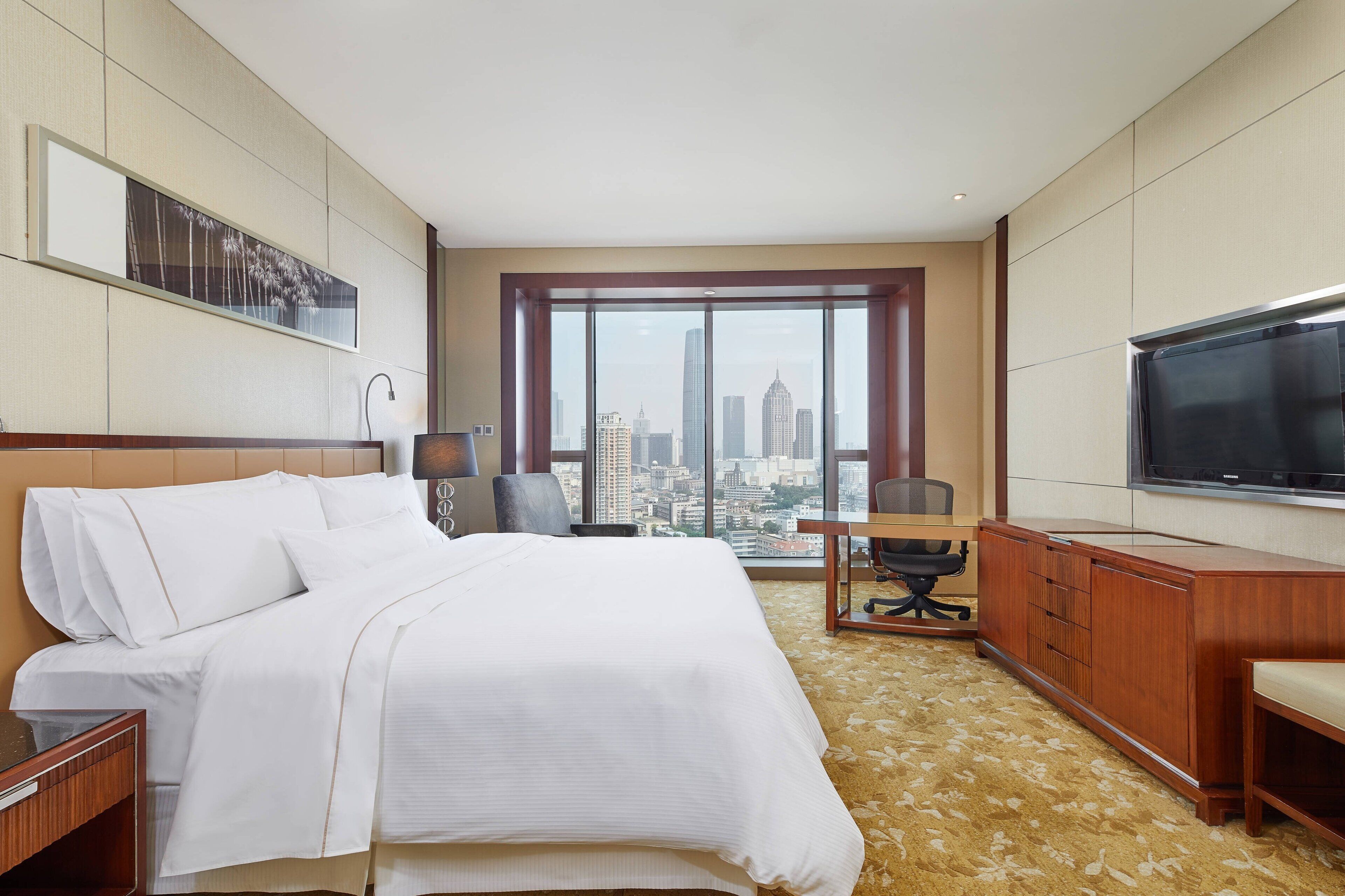 A hotel room with a large bed, a desk, a chair, a TV, and a view of the cityscape.