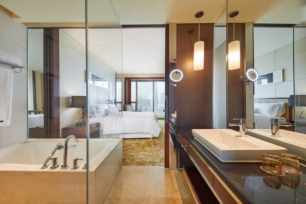 A modern hotel bathroom with a large bathtub, two sinks, and a city view through the glass door.
