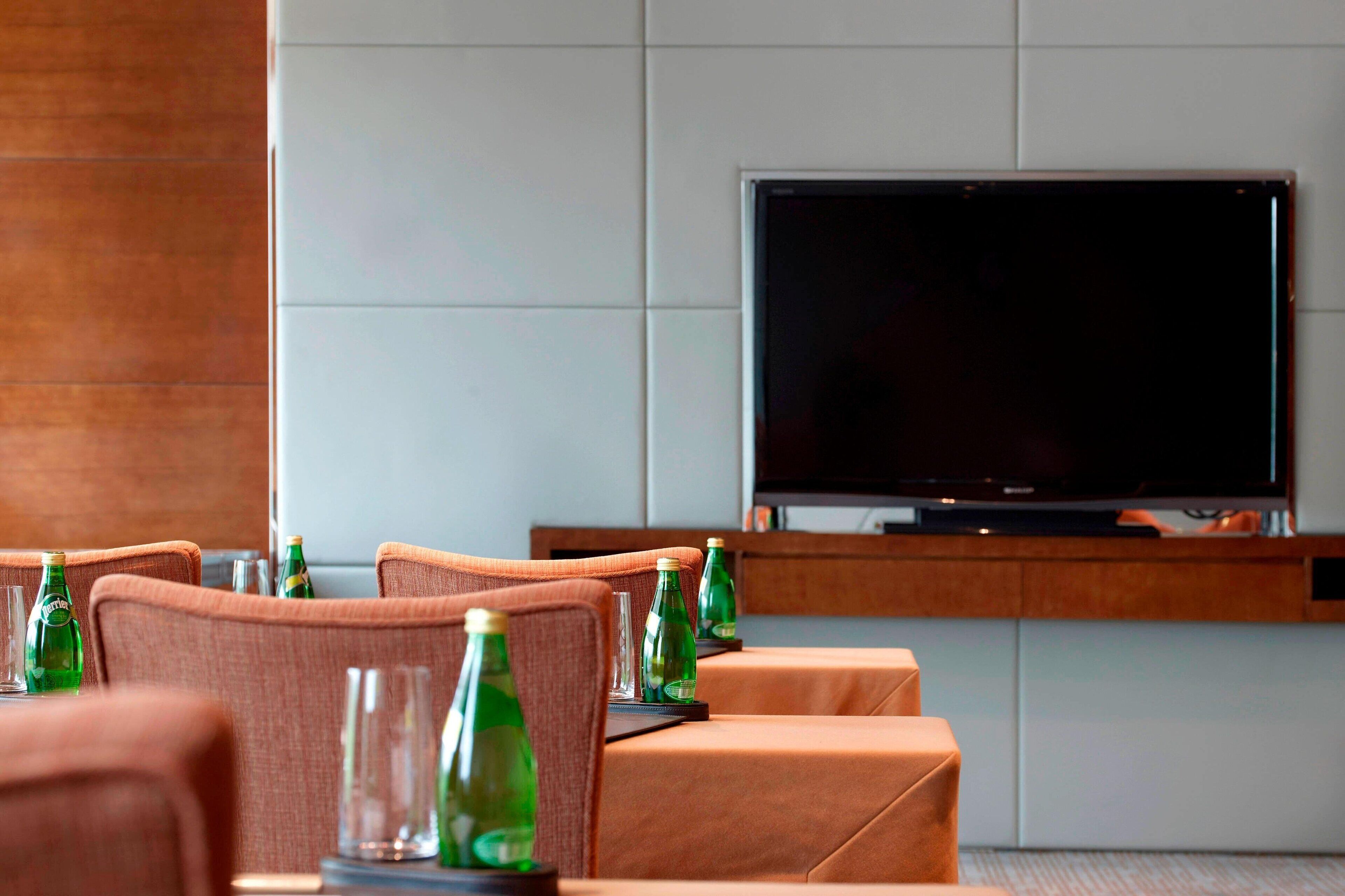 Habitació d'hotel moderna amb televisió de pantalla plana penjada a la paret, seients amb coixins i ampolles de vidre verd sobre la taula.