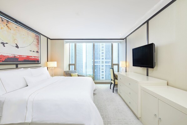 A hotel room with a large bed, a desk, a chair, a television, and a window with a city view.