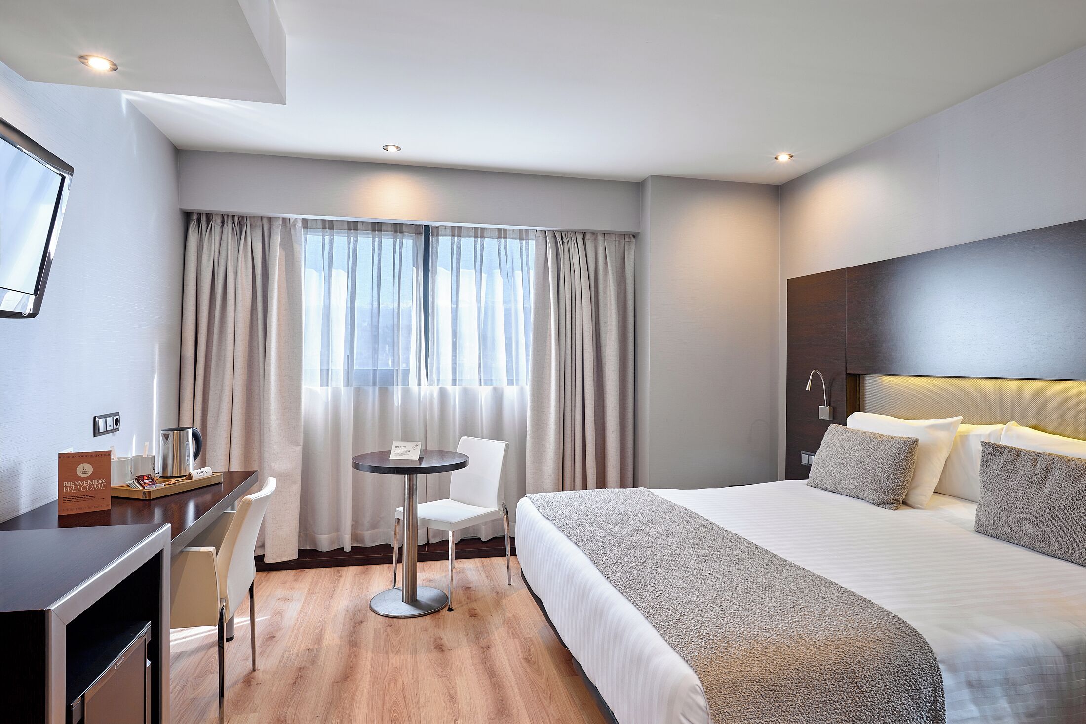 A hotel room with a bed, a desk with a chair, a small table, and a window with curtains.