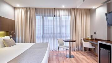 Habitació d'hotel amb llit, escriptori amb cadira, televisió i finestra amb cortines.