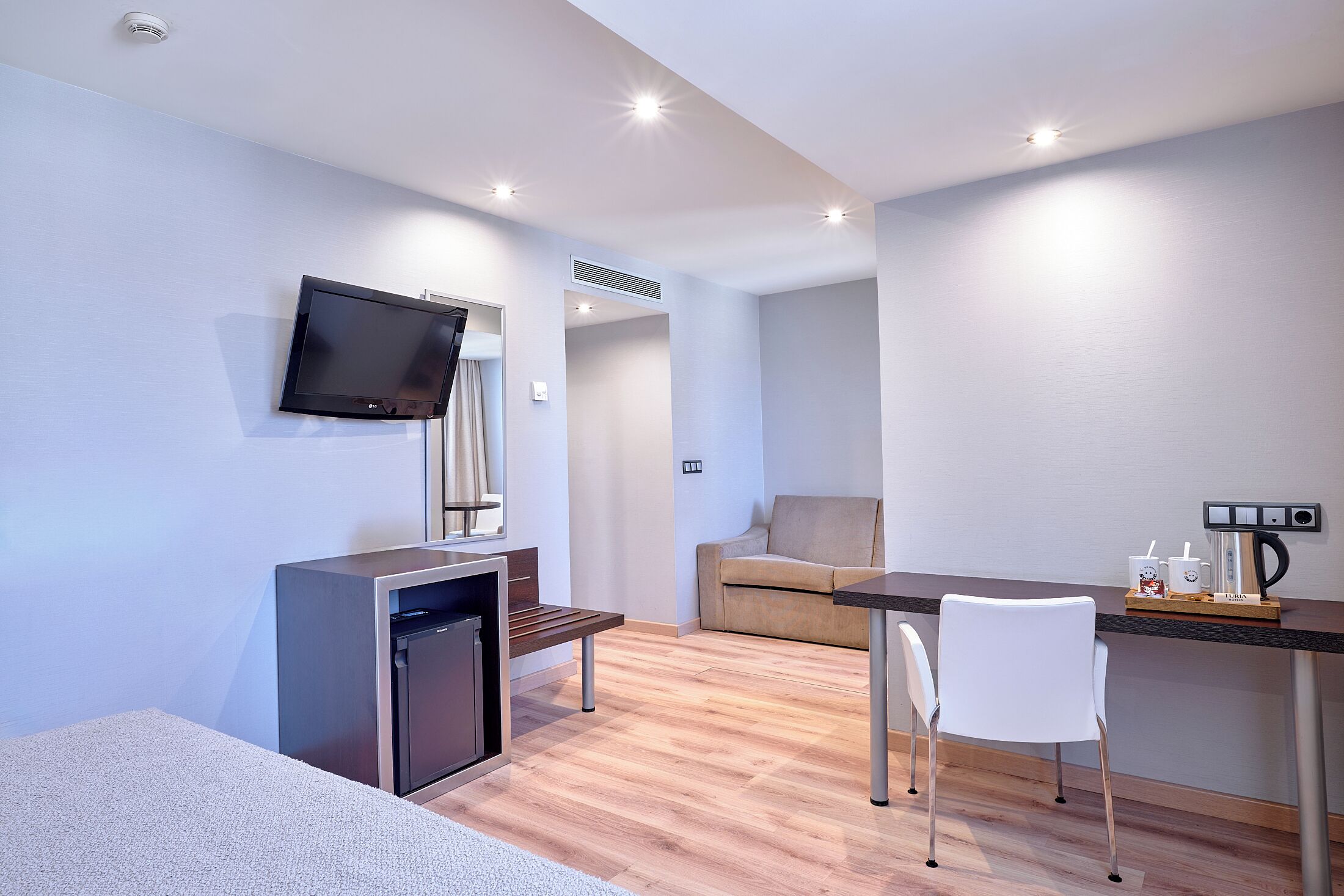 A modern room with a flat-screen TV, a small kitchenette, a sofa, a dining table with chairs, and a coffee machine.