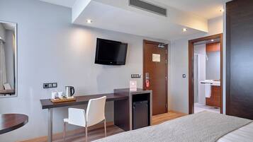 Habitación de hotel con cama, escritorio con silla, televisión, refrigerador pequeño y baño visible a través de una puerta abierta.