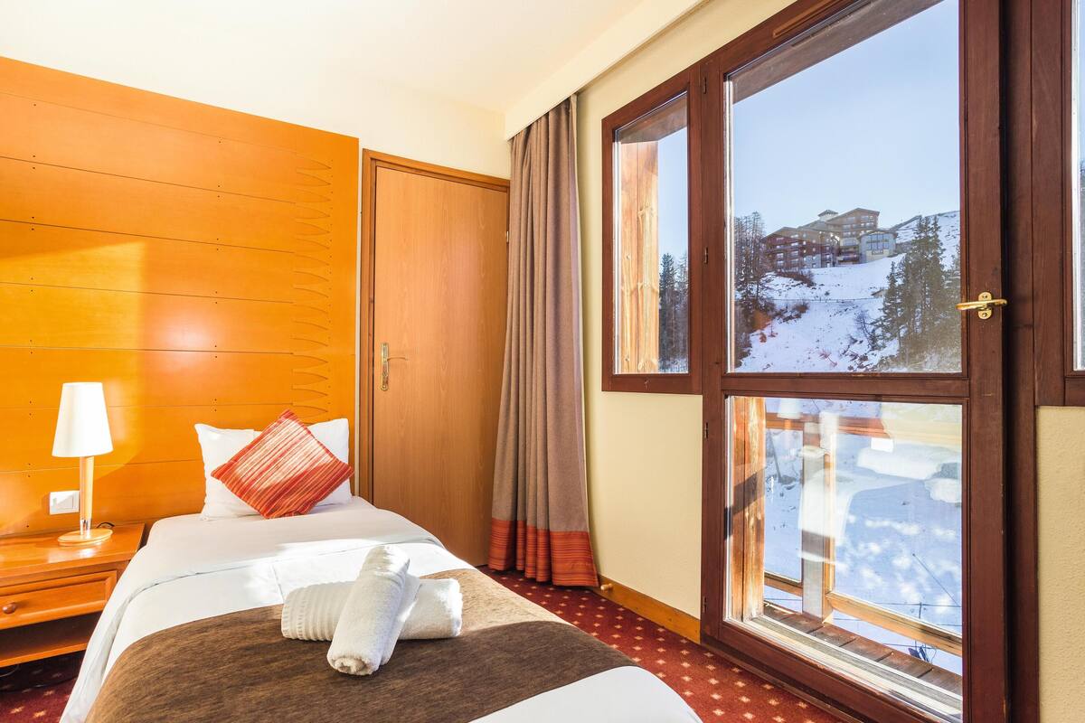 Ein Hotelzimmer mit großem Fenster, Holzwänden, einem Bett mit weißer Bettwäsche, einem roten Kissen und einem Nachttisch aus Holz mit einer Lampe.