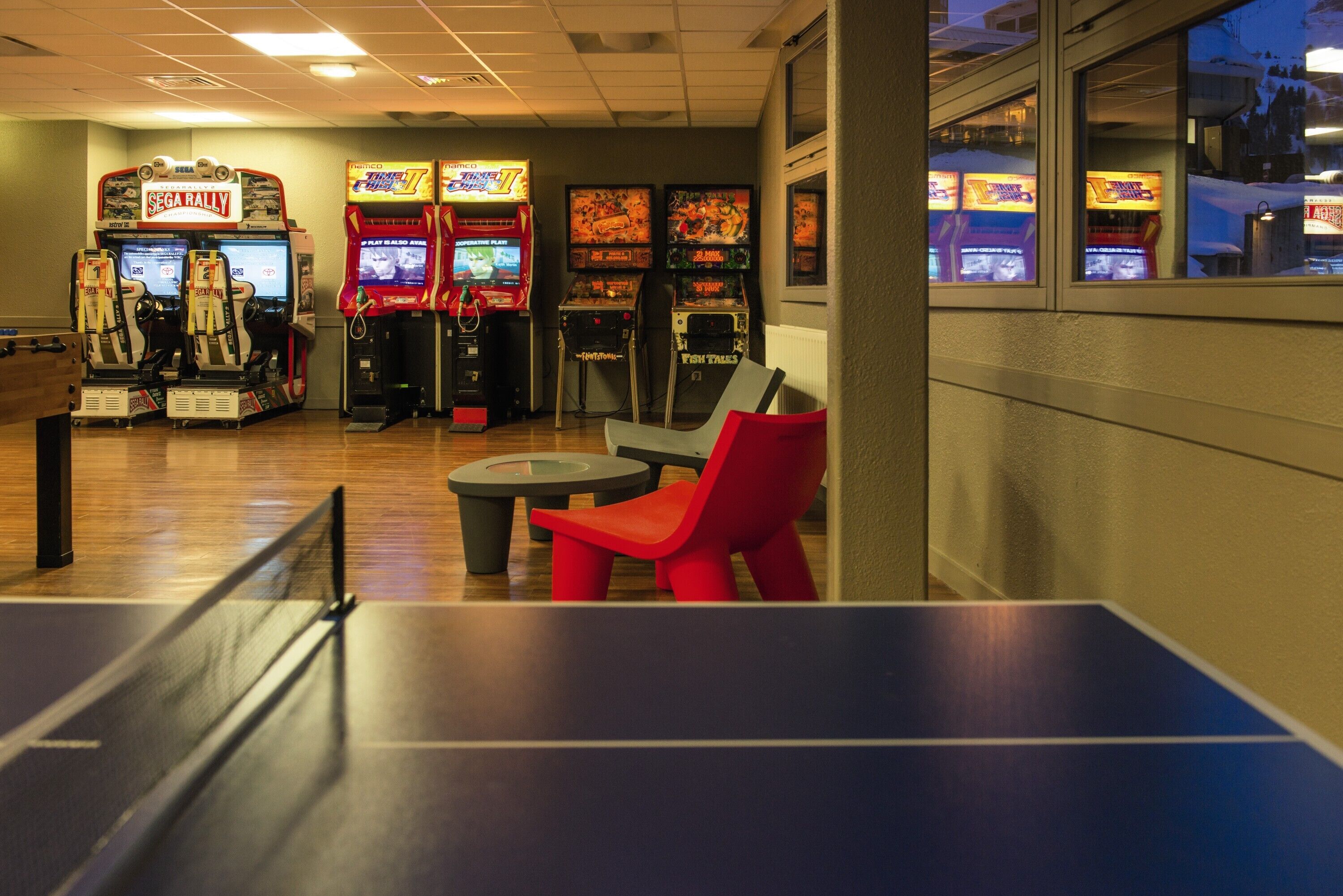 Una habitación con mesa de ping pong, sillas rojas y juegos de arcade, incluyendo SEGA RALLY y Fish Tales.