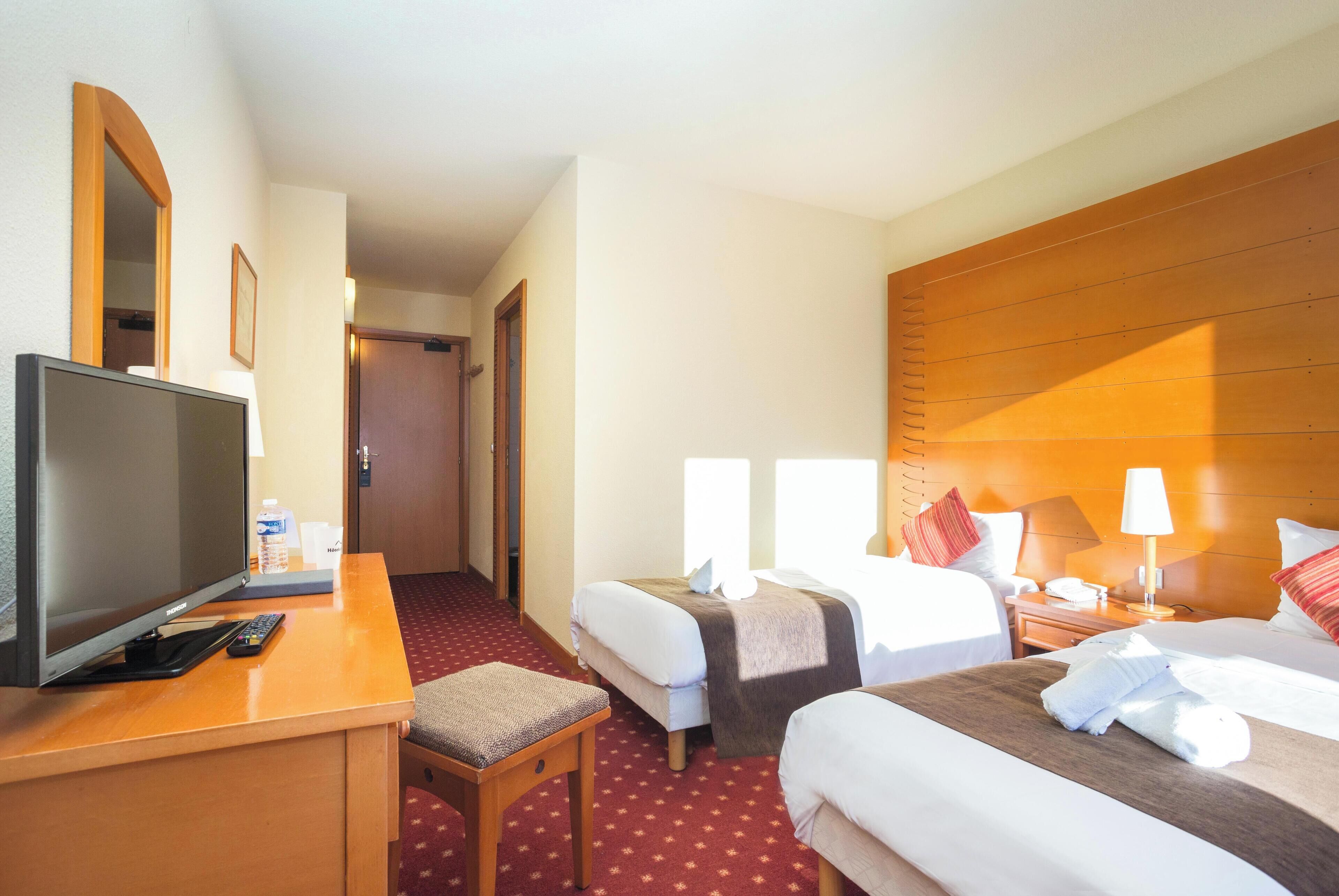 A hotel room with two beds, a desk with a TV, a chair, and a wooden headboard.