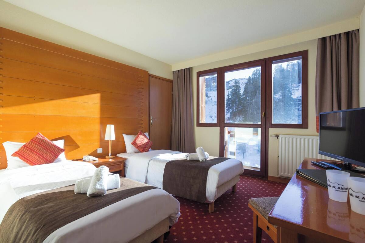 Ein Hotelzimmer mit zwei Betten, einem Schreibtisch mit Fernseher und einem Fenster mit Blick auf schneebedeckte Bäume.