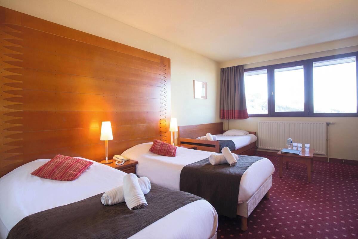 Ein Hotelzimmer mit zwei Betten, einem Holzkopfstück, einem Fenster mit Vorhängen, einem kleinen Tisch mit einer Lampe und einem Heizkörper.